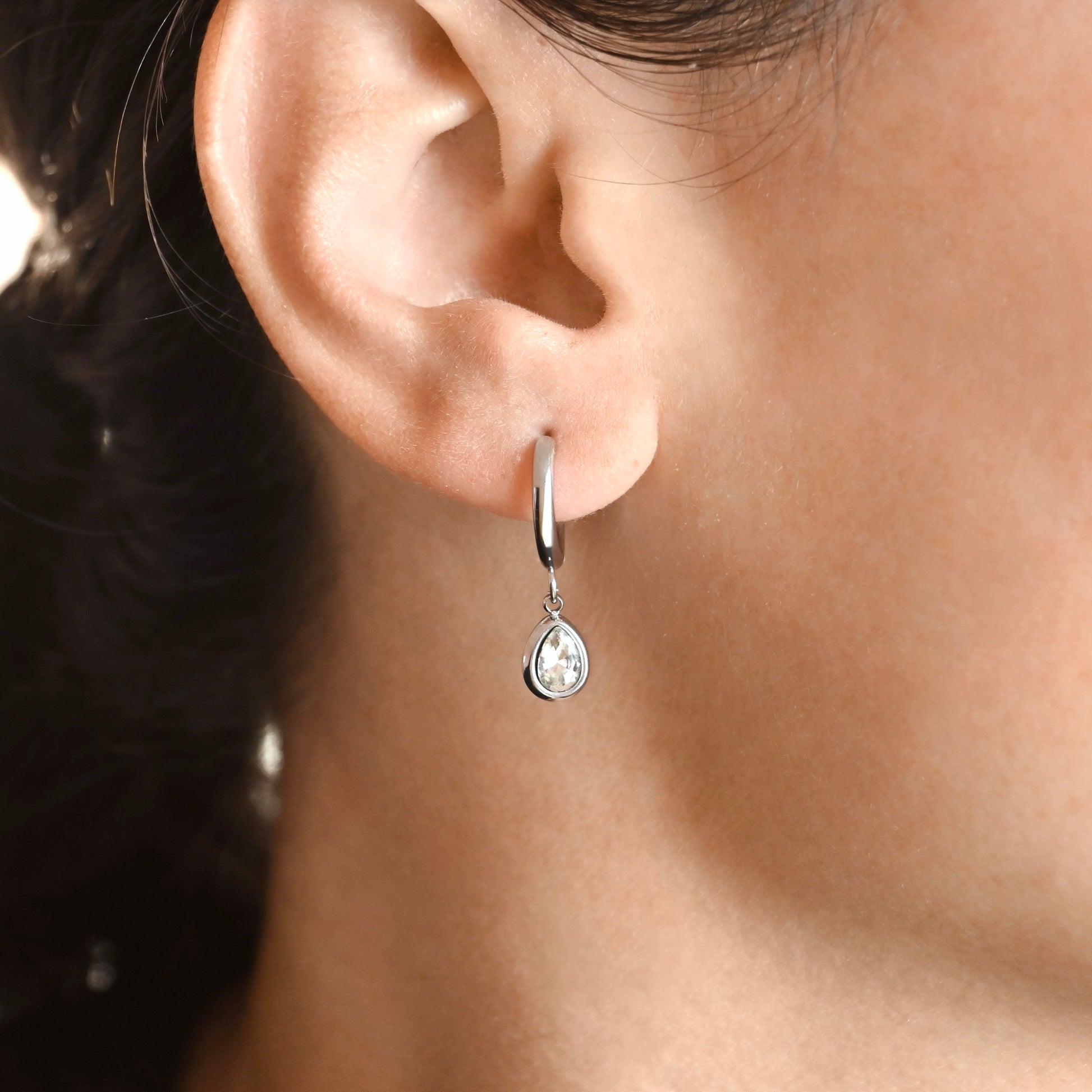 A close-up of a person's ear wearing a small silver huggie hoop earring with a teardrop-shaped clear gemstone dangle.
