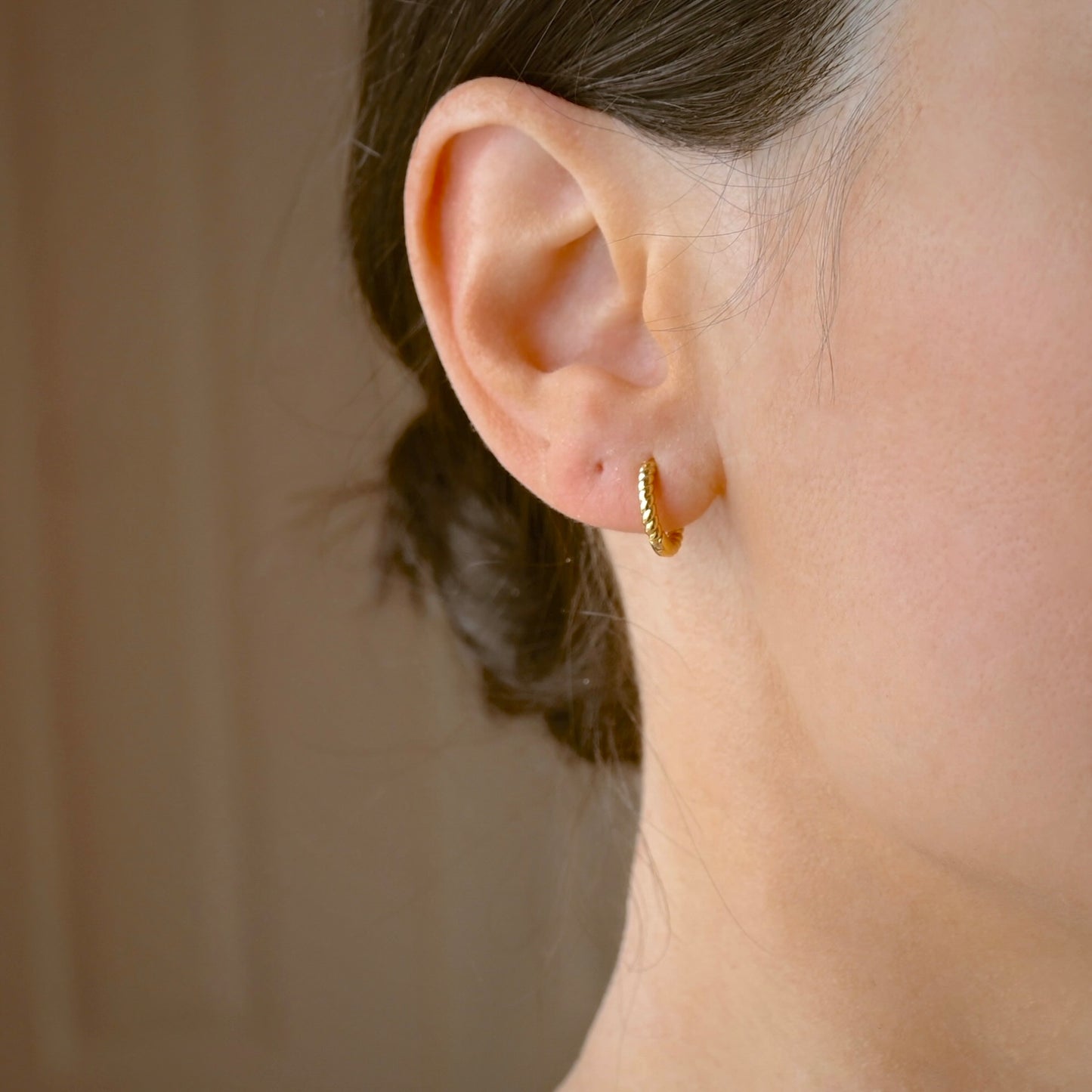 A close-up of a person's ear wearing a small gold twisted huggie hoop earring, with the hair pulled back and a soft neutral background.