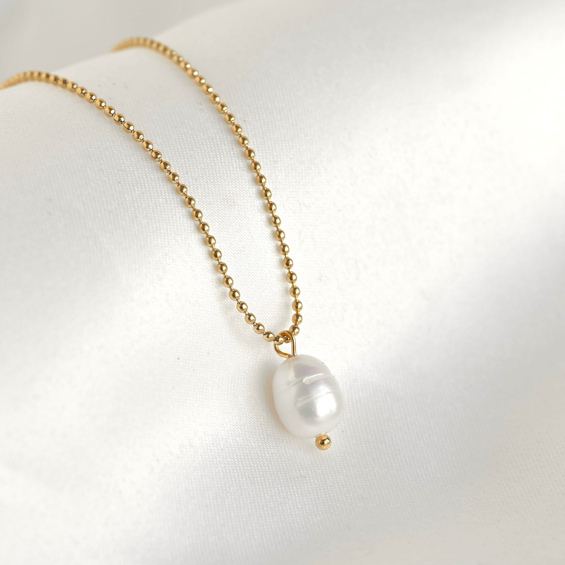 A delicate gold ball-chain necklace featuring a single white baroque pearl pendant with a small gold bead accent below it, displayed on a soft white fabric surface.