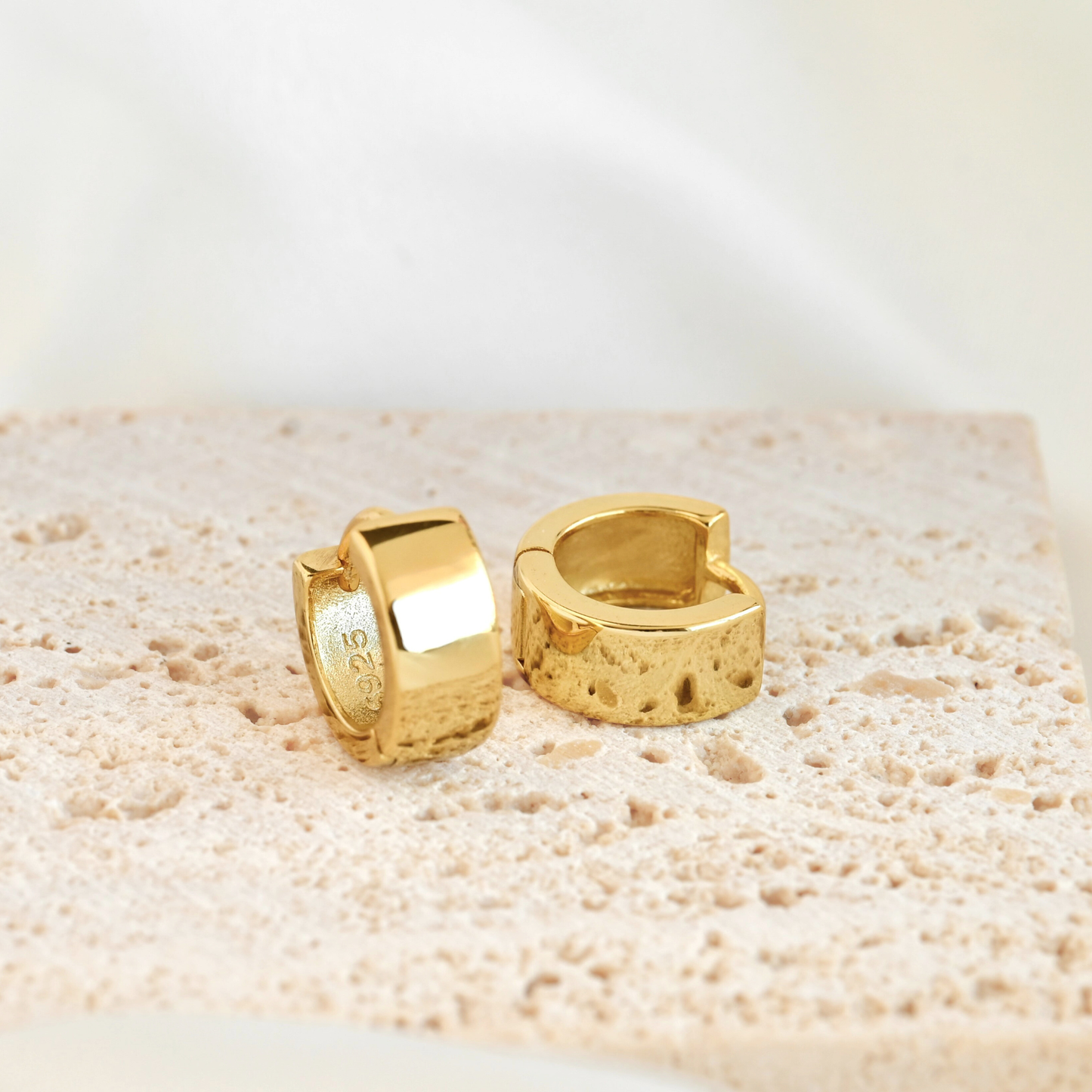 A pair of thick gold huggie hoop earrings displayed on a beige stone slab.