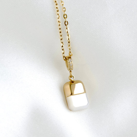 A gold chain necklace with a capsule-shaped pendant, featuring a polished gold top and bottom half separated by a white center, displayed on a white satin fabric background.