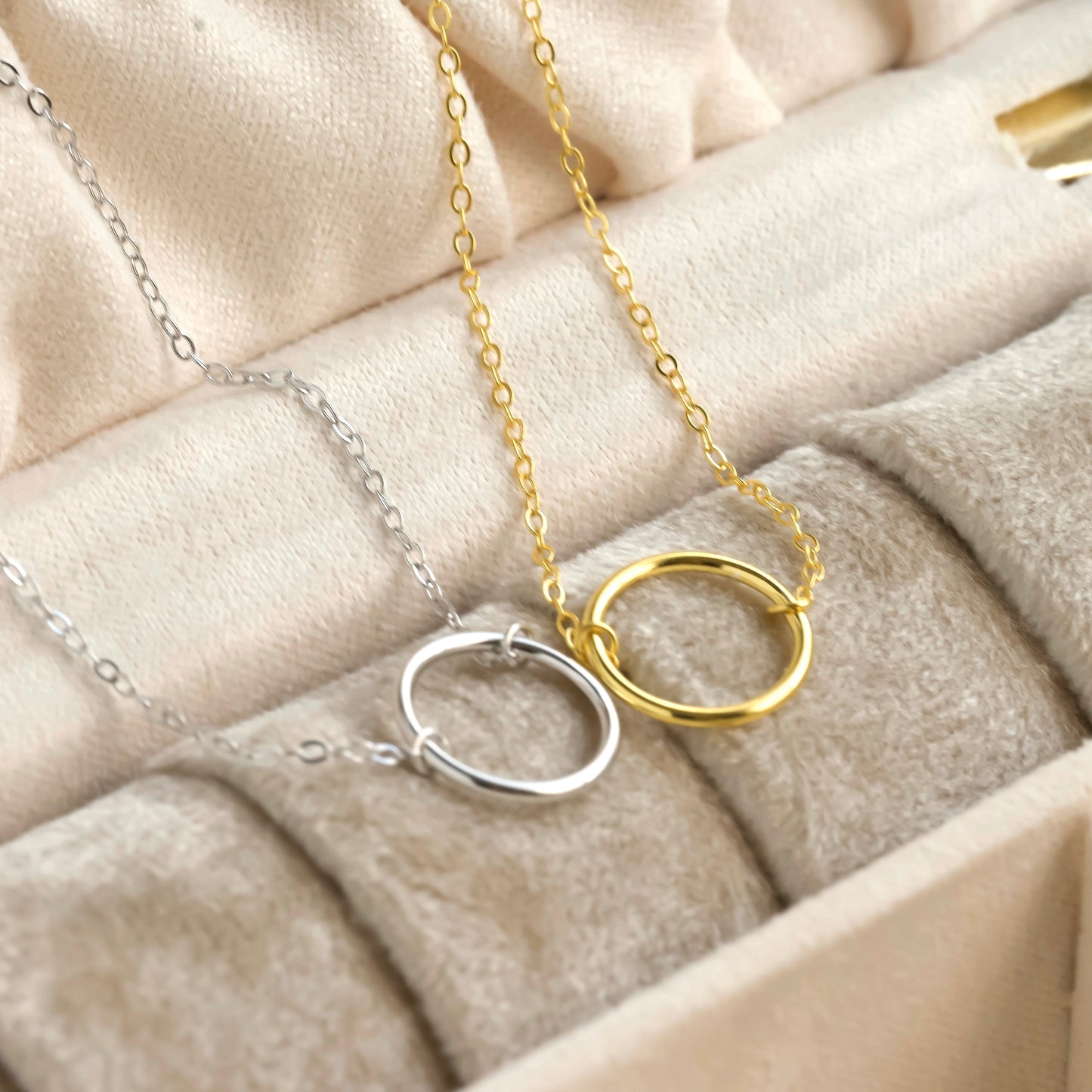 Two eternity necklaces, one gold and one silver, on a soft beige fabric background.