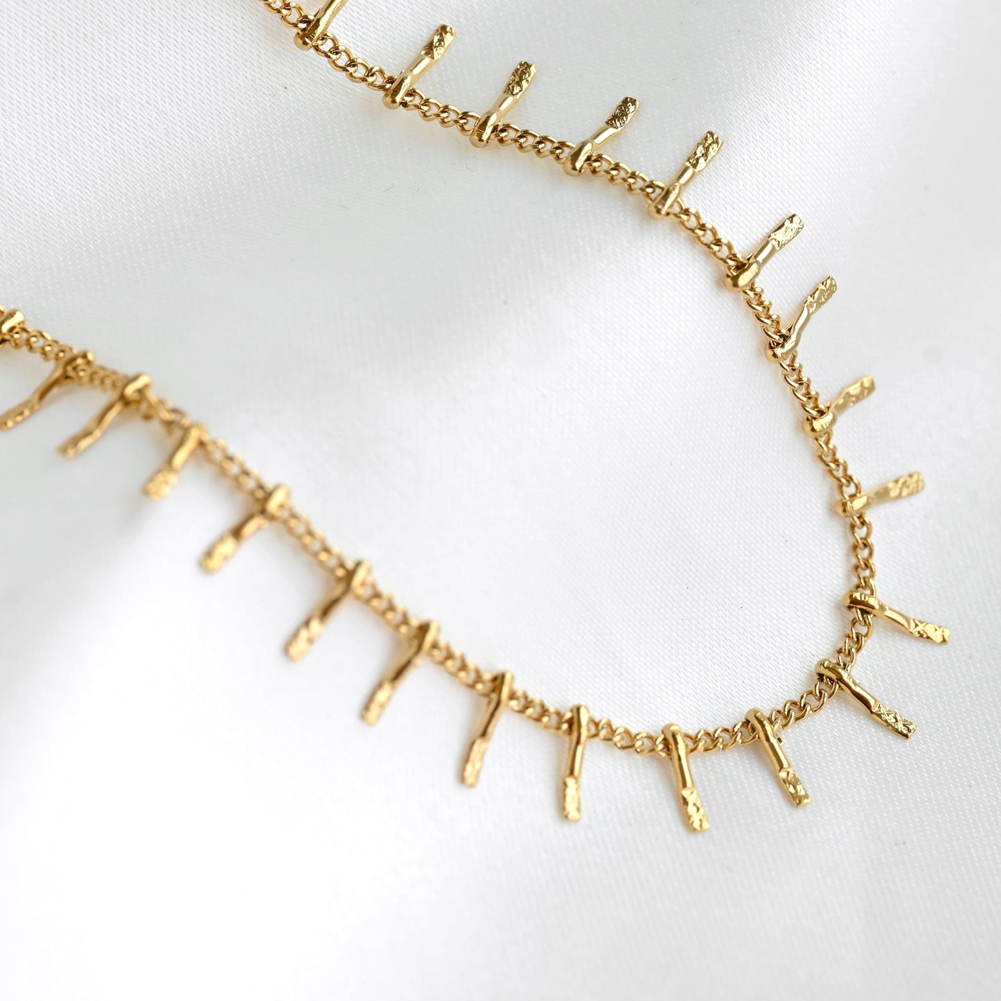 A gold chain necklace featuring small, textured vertical spikes along the front, displayed on a soft white fabric background.