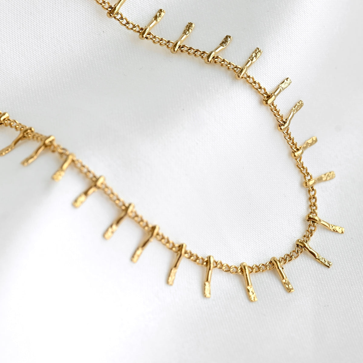 A gold chain necklace featuring small, textured vertical spikes along the front, displayed on a soft white fabric background.