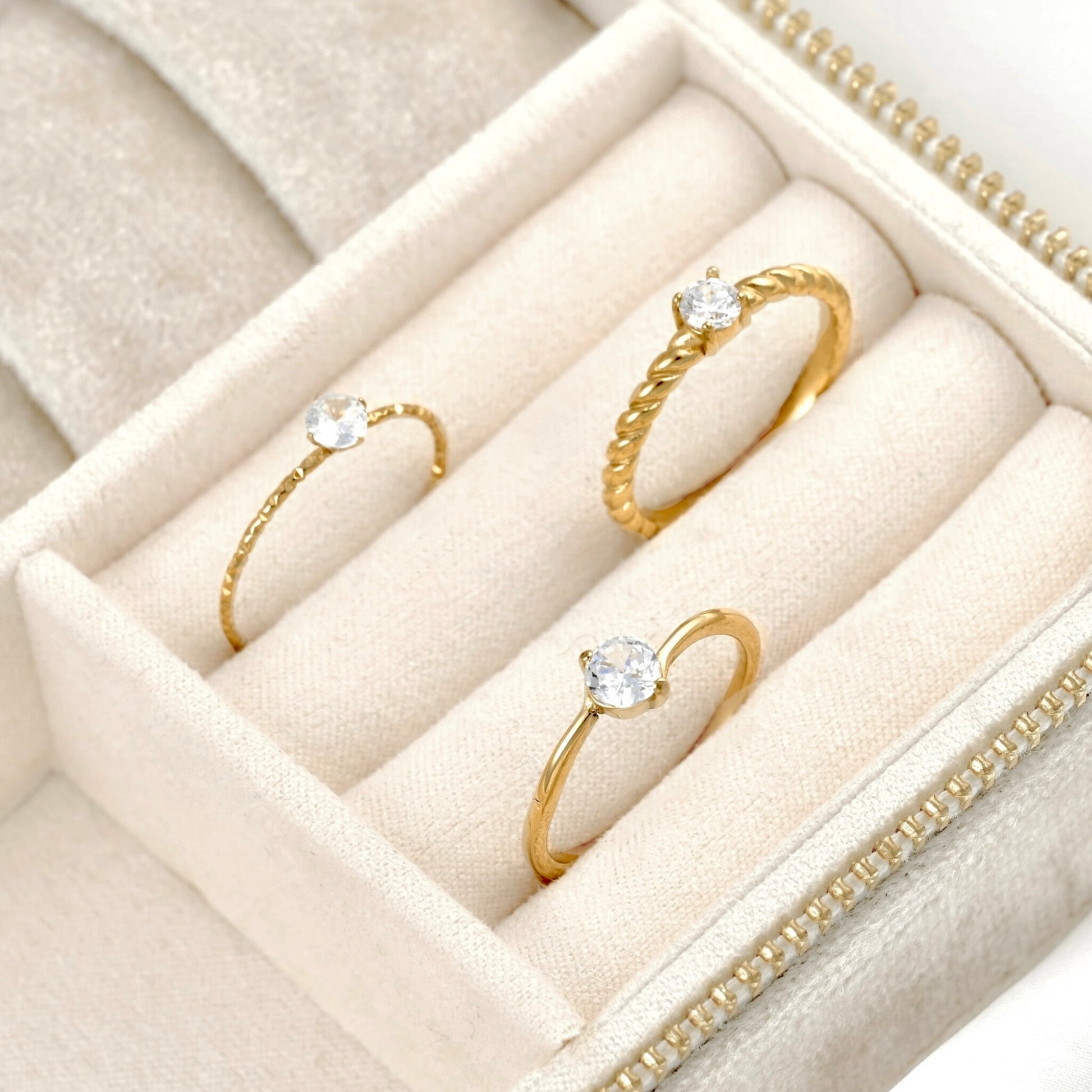 Three gold rings with clear gemstones displayed in a cream-colored velvet jewelry box.