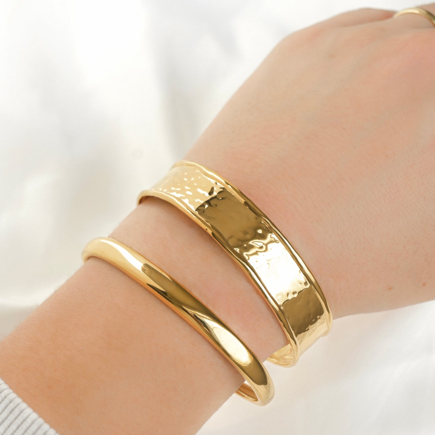 A close-up of a person's wrist wearing two stacked gold bangle bracelets, including one wide hammered-texture cuff, and a slim doff cuff against a soft white background.