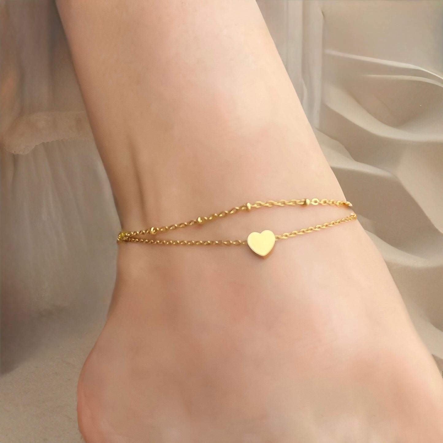 Layered gold anklets on a person's ankle with a heart-shaped charm against a neutral background