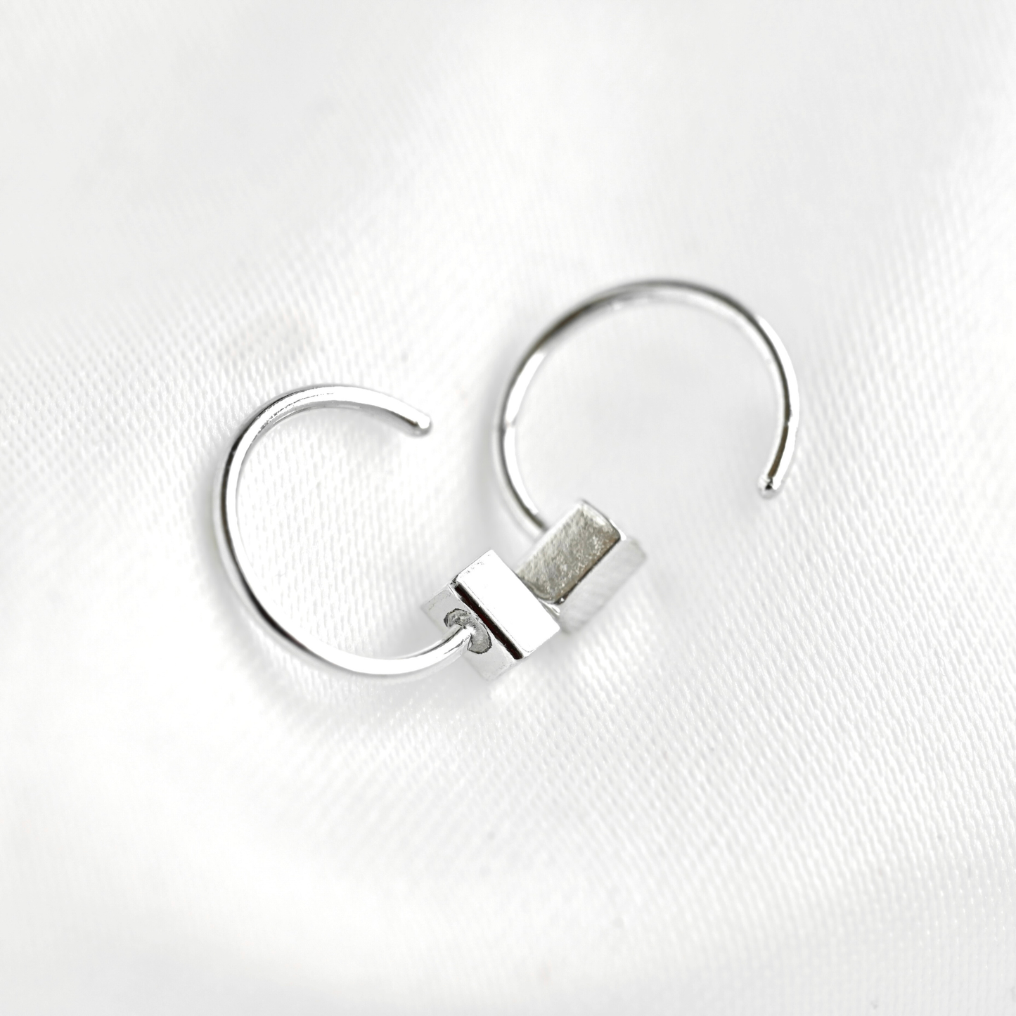 A pair of silver-toned open hoop earrings with a small rectangular bar detail, displayed on a textured white fabric background.