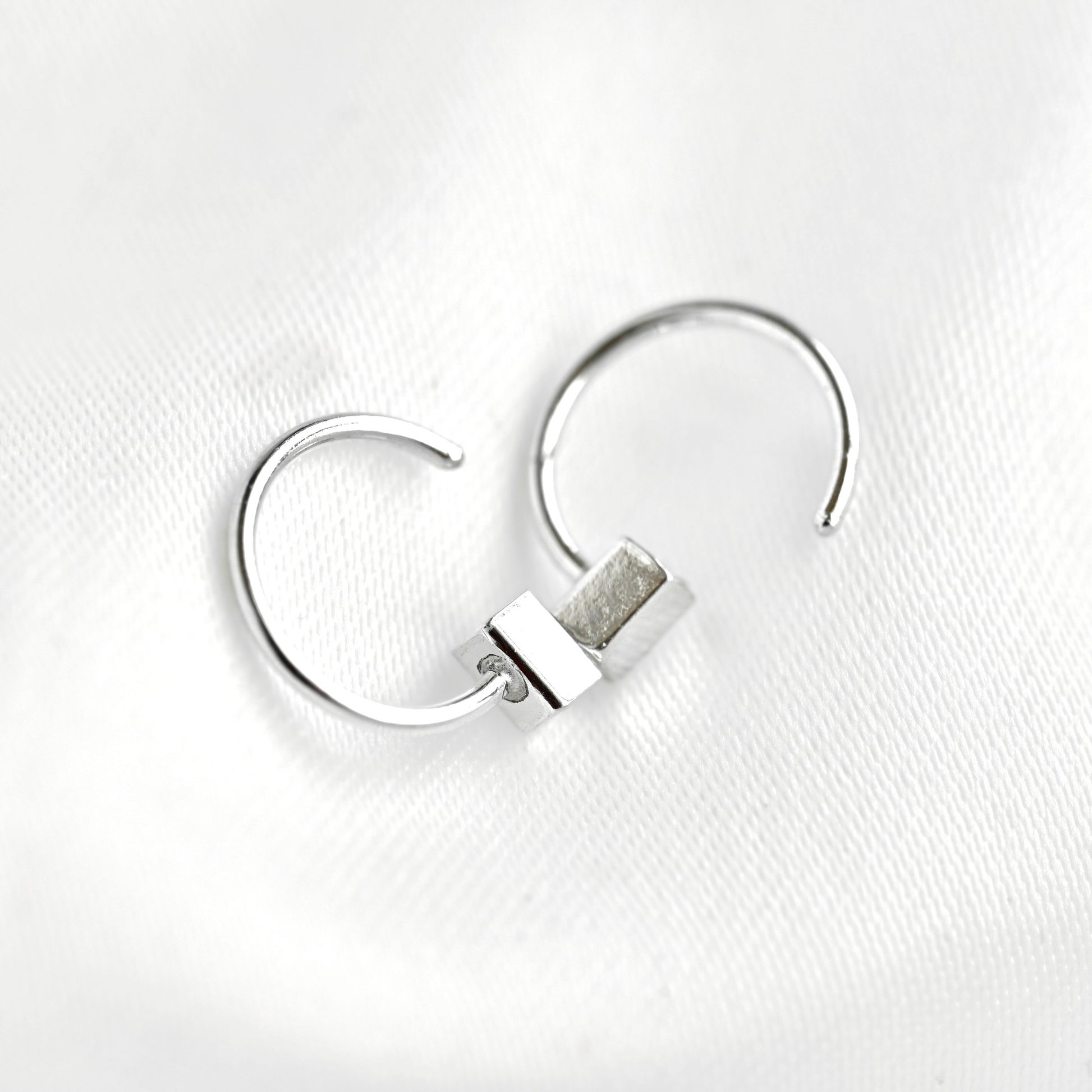A pair of silver-toned open hoop earrings with a small rectangular bar detail, displayed on a textured white fabric background.