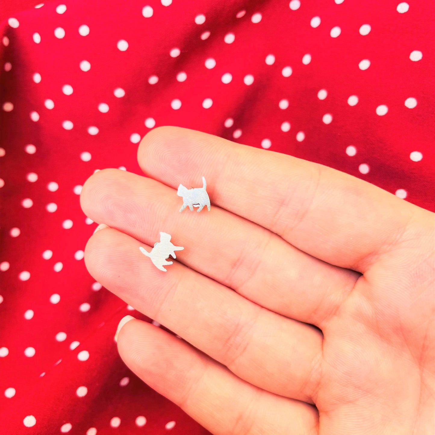 Bright silver cat-shaped stud earrings held between fingers against a red polka dot background