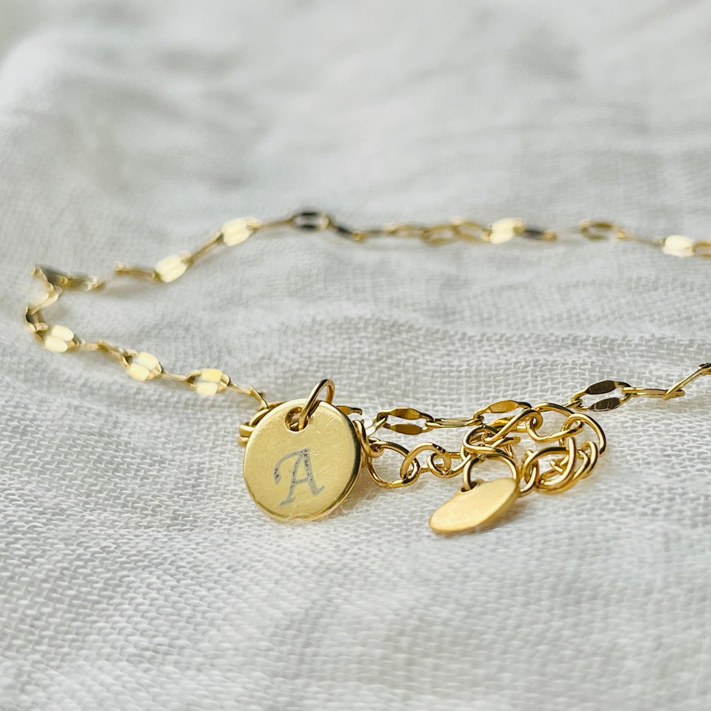 Gold sequin anklet with  a letter charm on a light gray fabric background