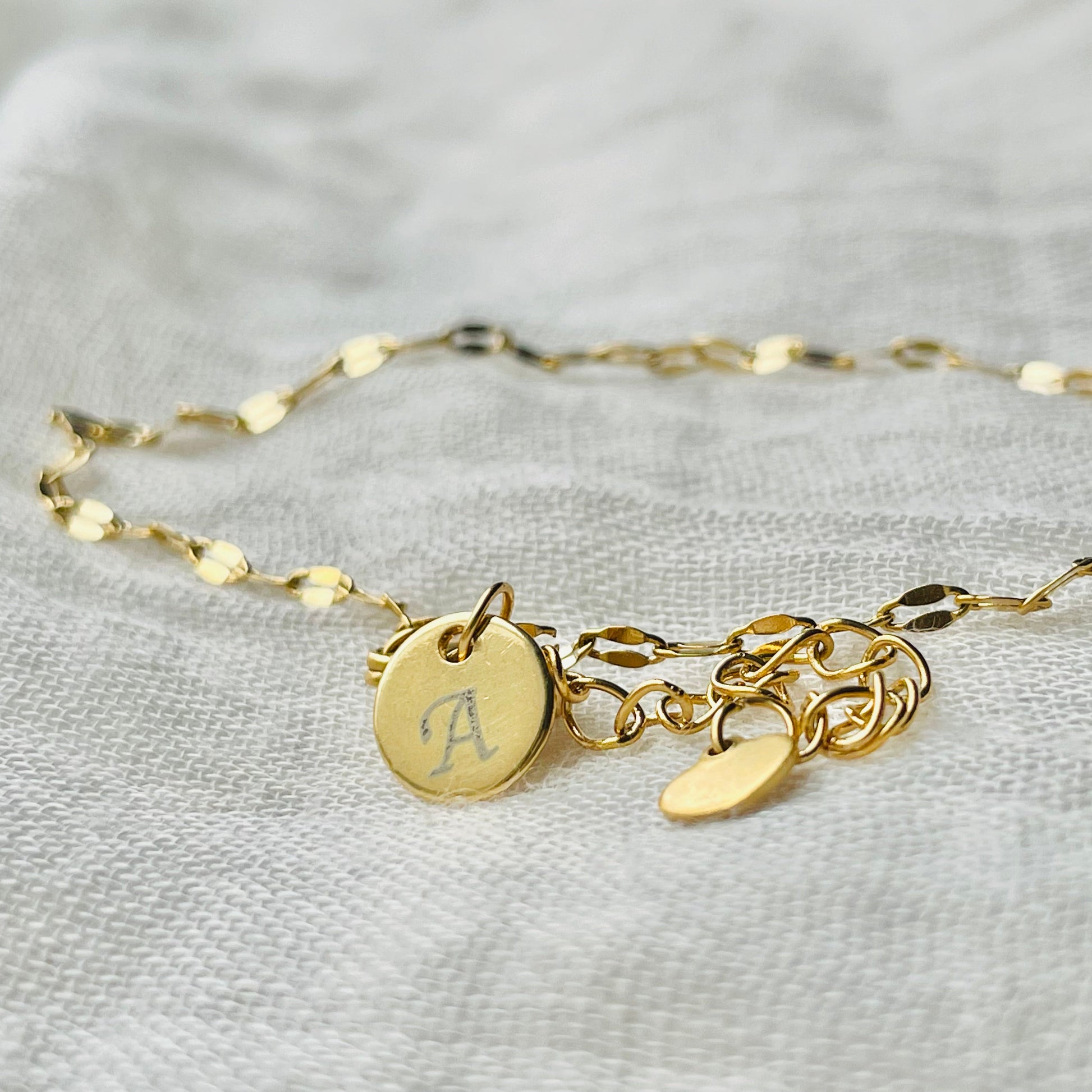 Gold sequin anklet with  a letter charm on a light gray fabric background