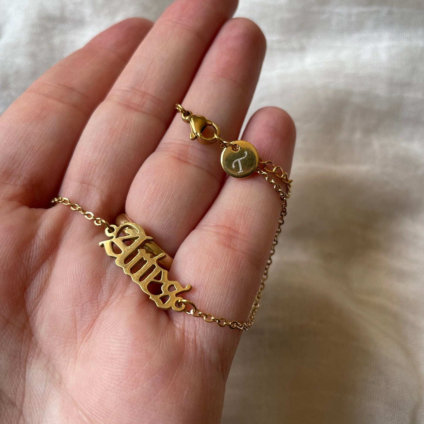 Gold anklet with a zodiac charm and engraved initial disc at the clasp being held on a hand against a white fabric background