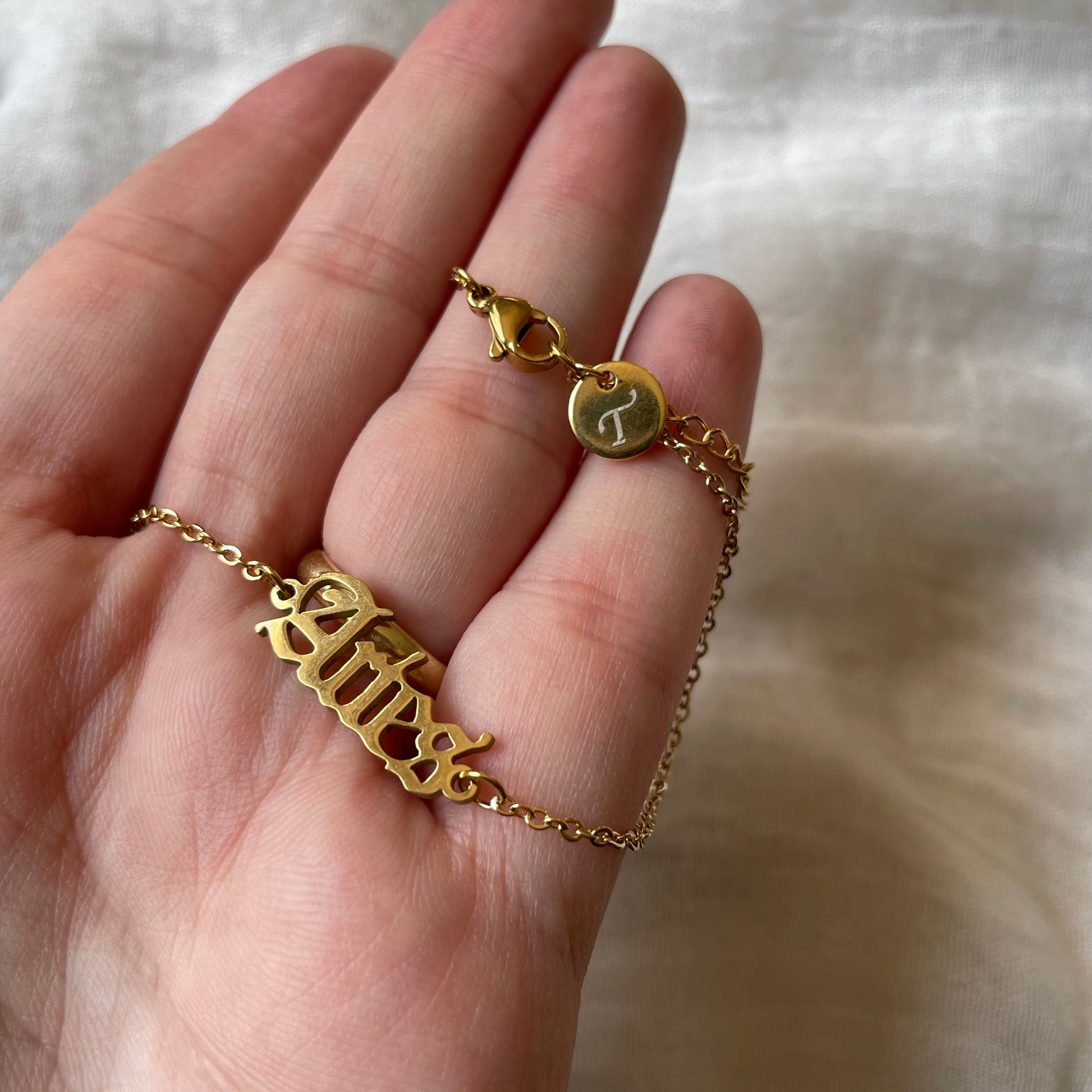 Gold anklet with a zodiac charm and engraved initial disc at the clasp being held on a hand against a white fabric background