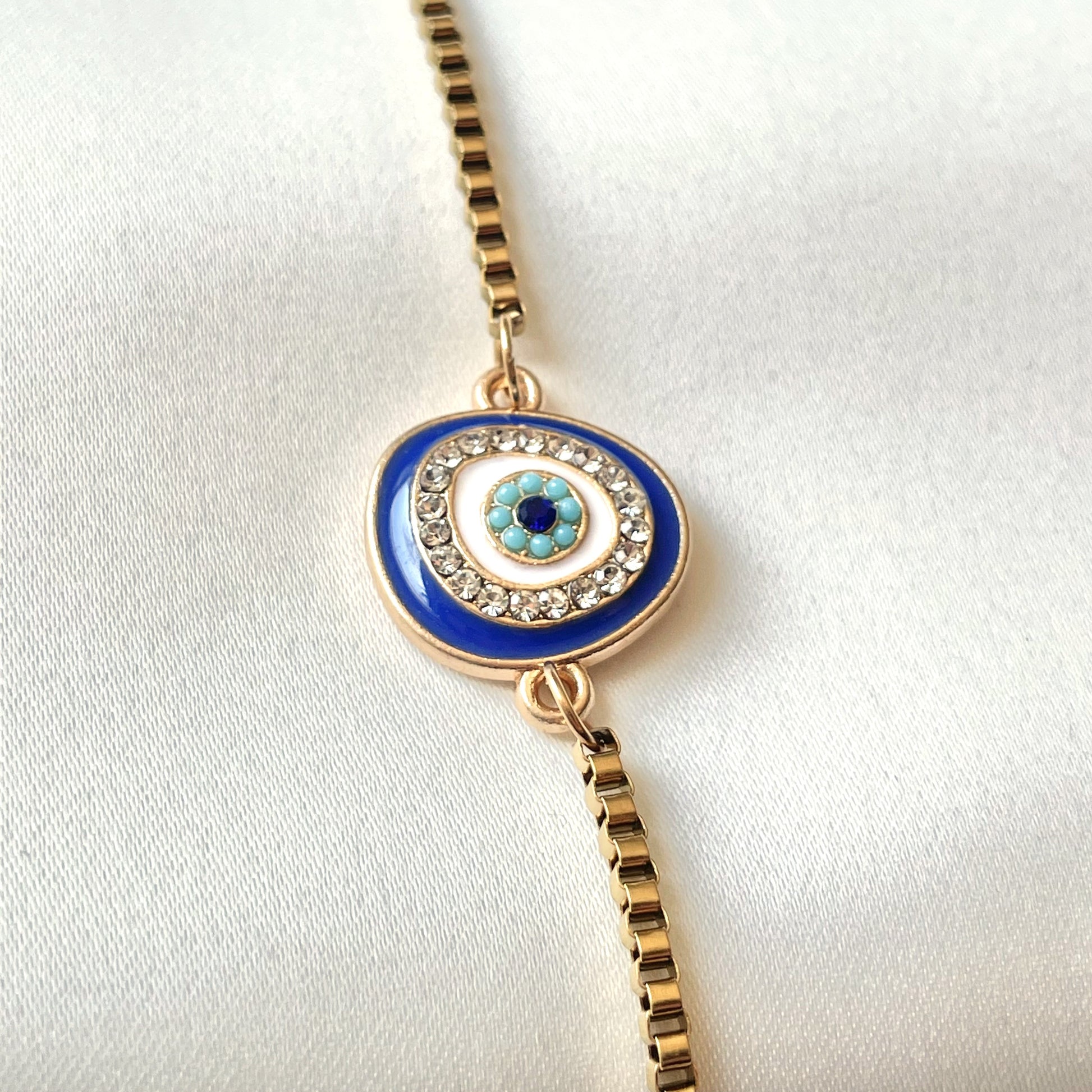 Close-up view of the organic shaped evil eye charm featuring dark blue enamel, a row of clear stones, white enamel, turquoise beads and finally a sapphire colored stone centerpiece.