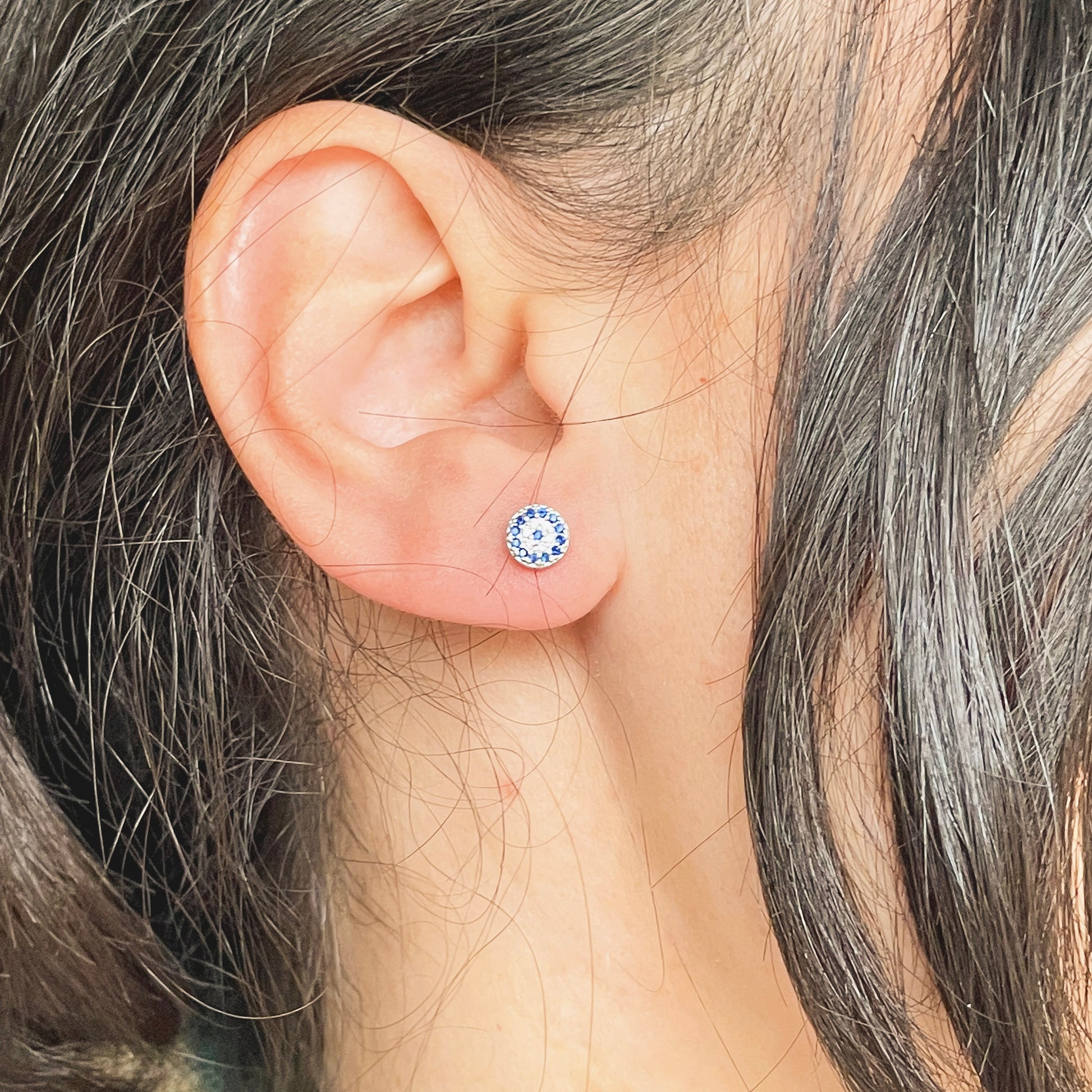 Close-up of an ear wearing small round silver stud earrings with blue and clear CZ stones