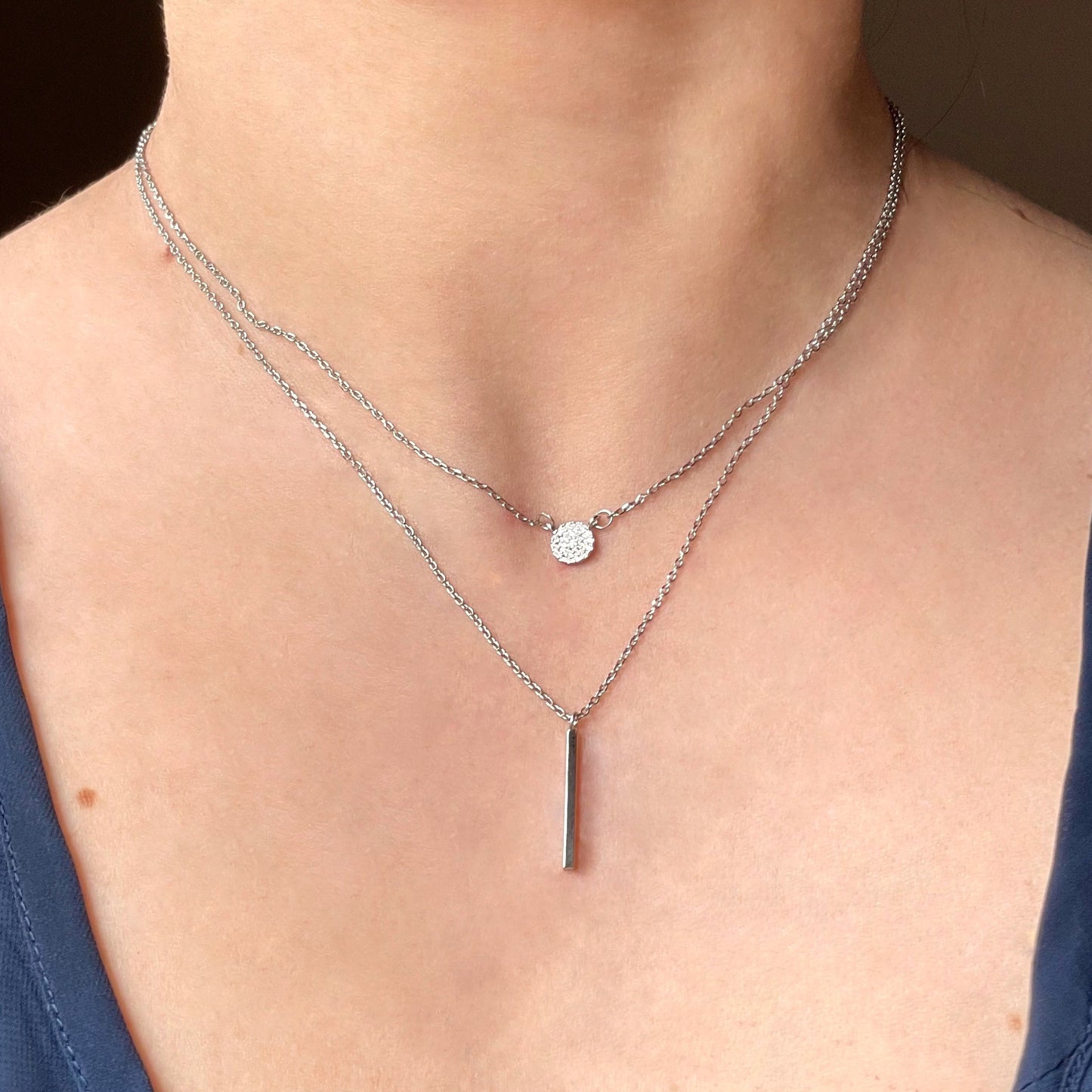 Close-up of a person wearing double layered silver necklaces with a hammered disc charm and vertical bar pendant with a plain background