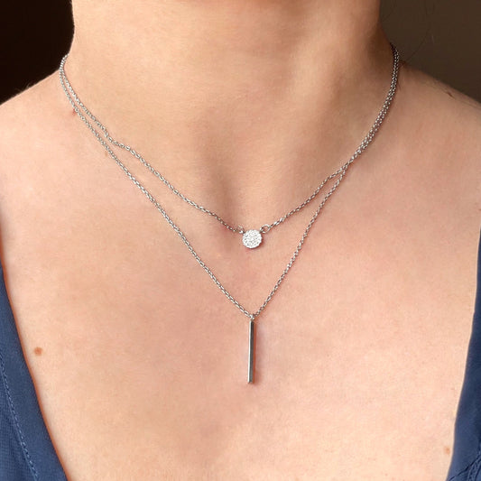 Close-up of a person wearing double layered silver necklaces with a hammered disc charm and vertical bar pendant with a plain background