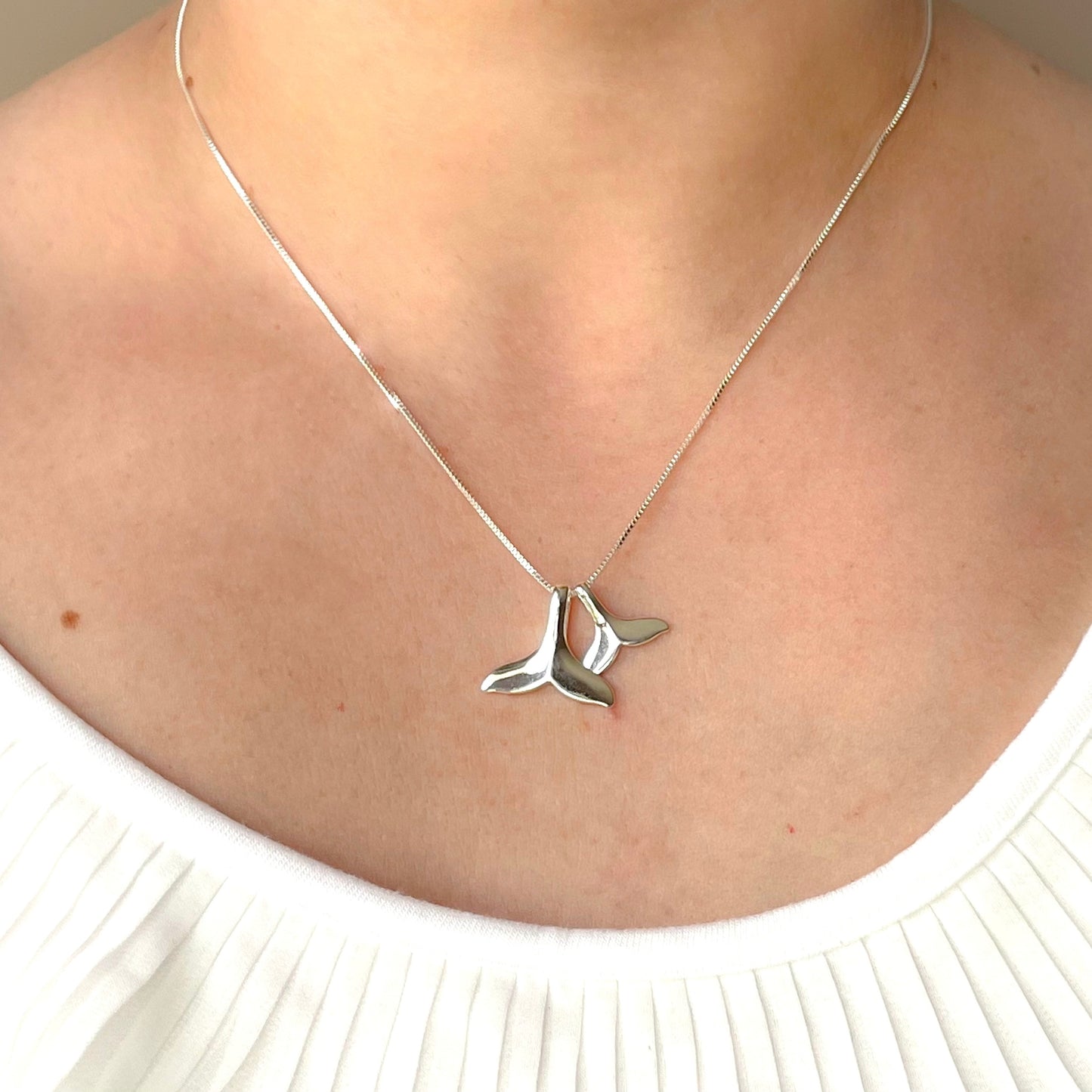 Silver whale tail necklace with two pendants, one big and one small, on a person wearing a white top 