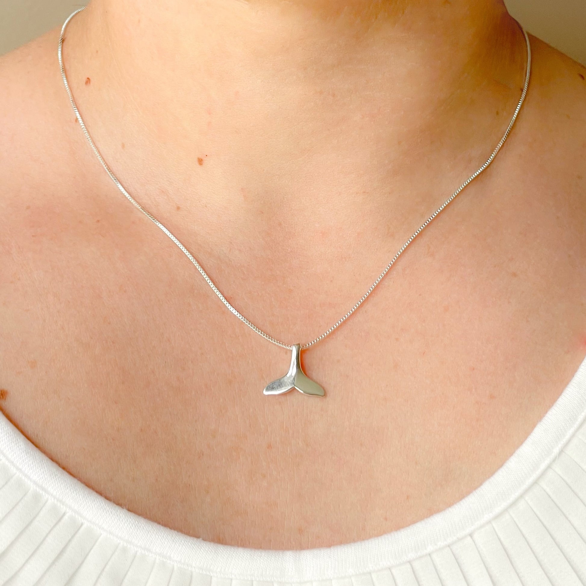 Close-up front view of silver necklace with a whale tail pendant on a person wearing a white top.