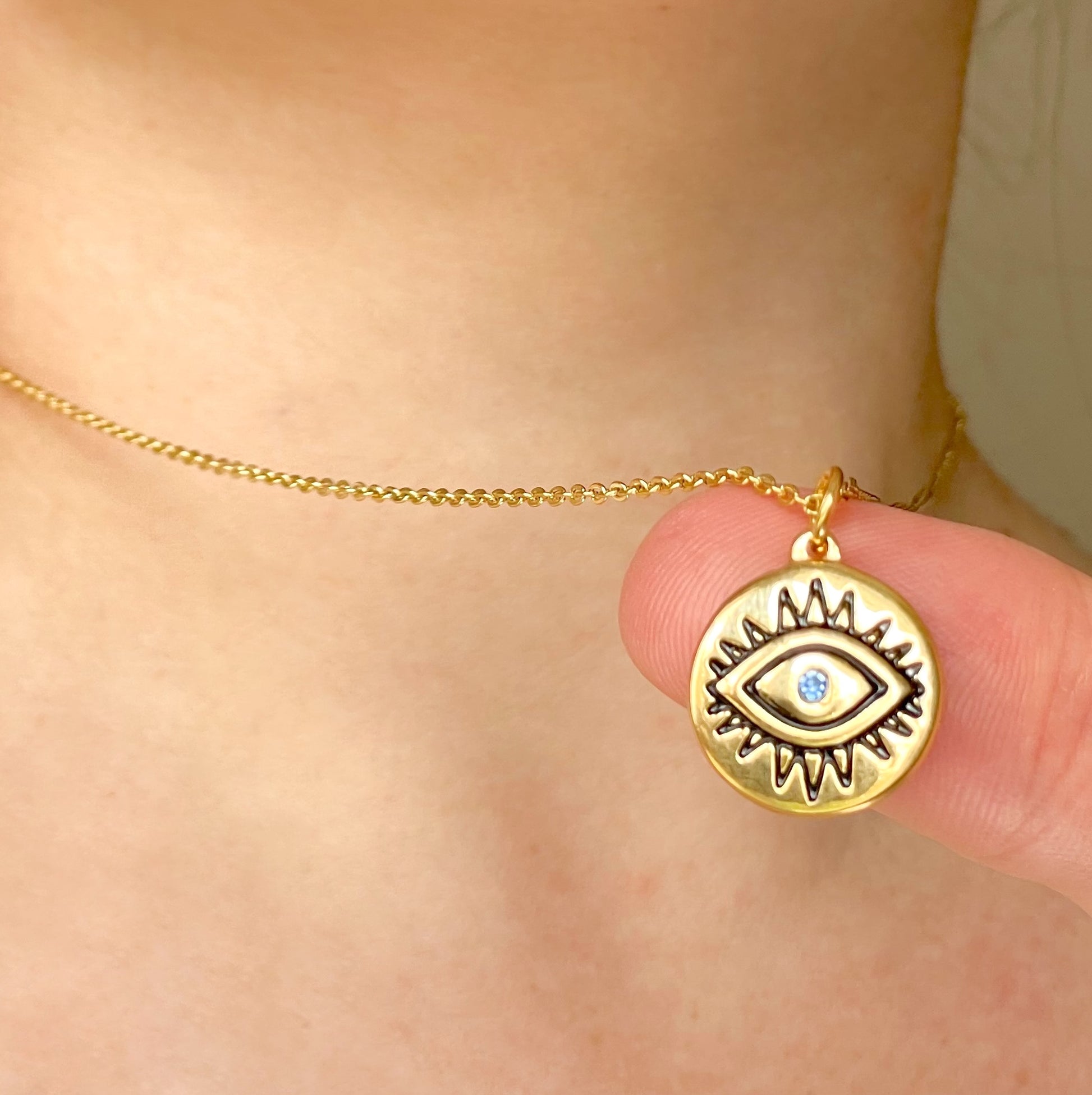 Gold necklace with an eye-shaped pendant and cable chain held by a hand against a neutral background