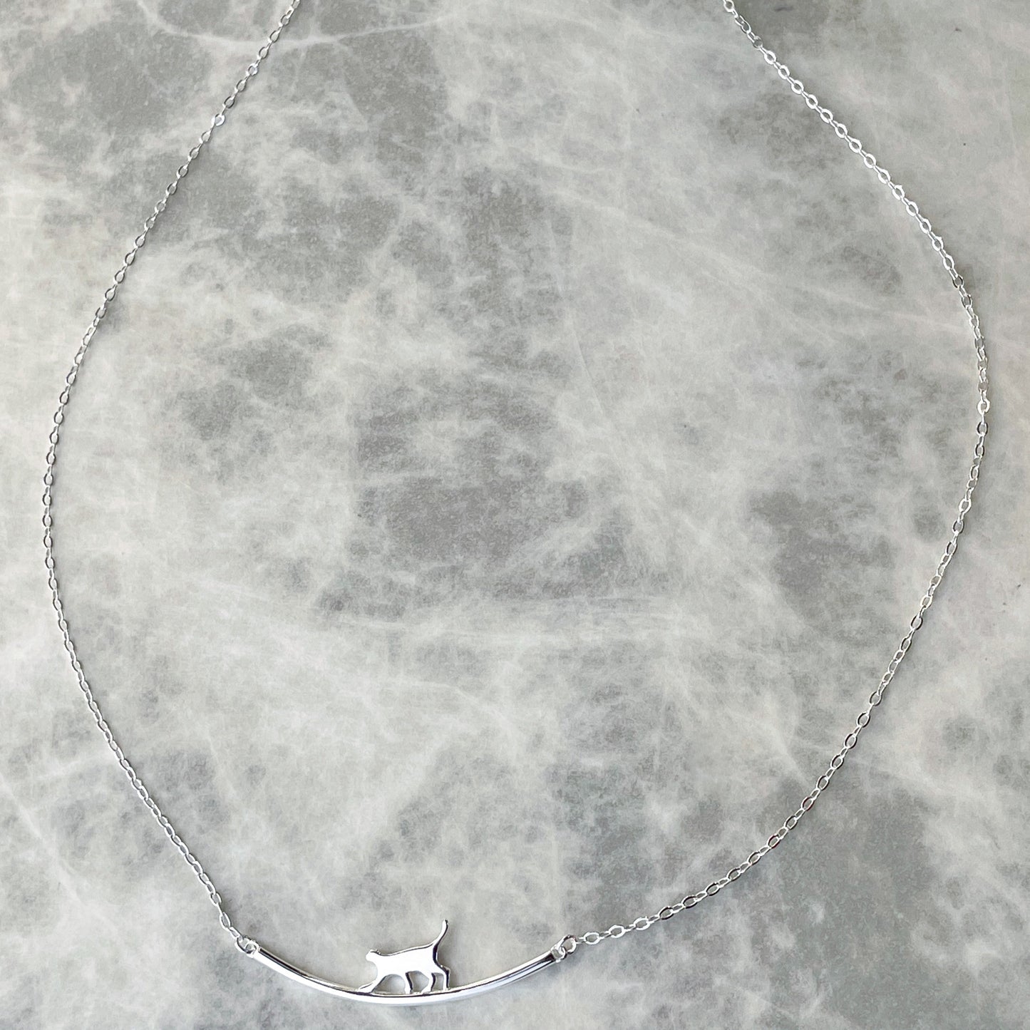 Wide angled view of silver necklace with a long curved bar pendant with a walking  cat silhouette on a gray marble surface