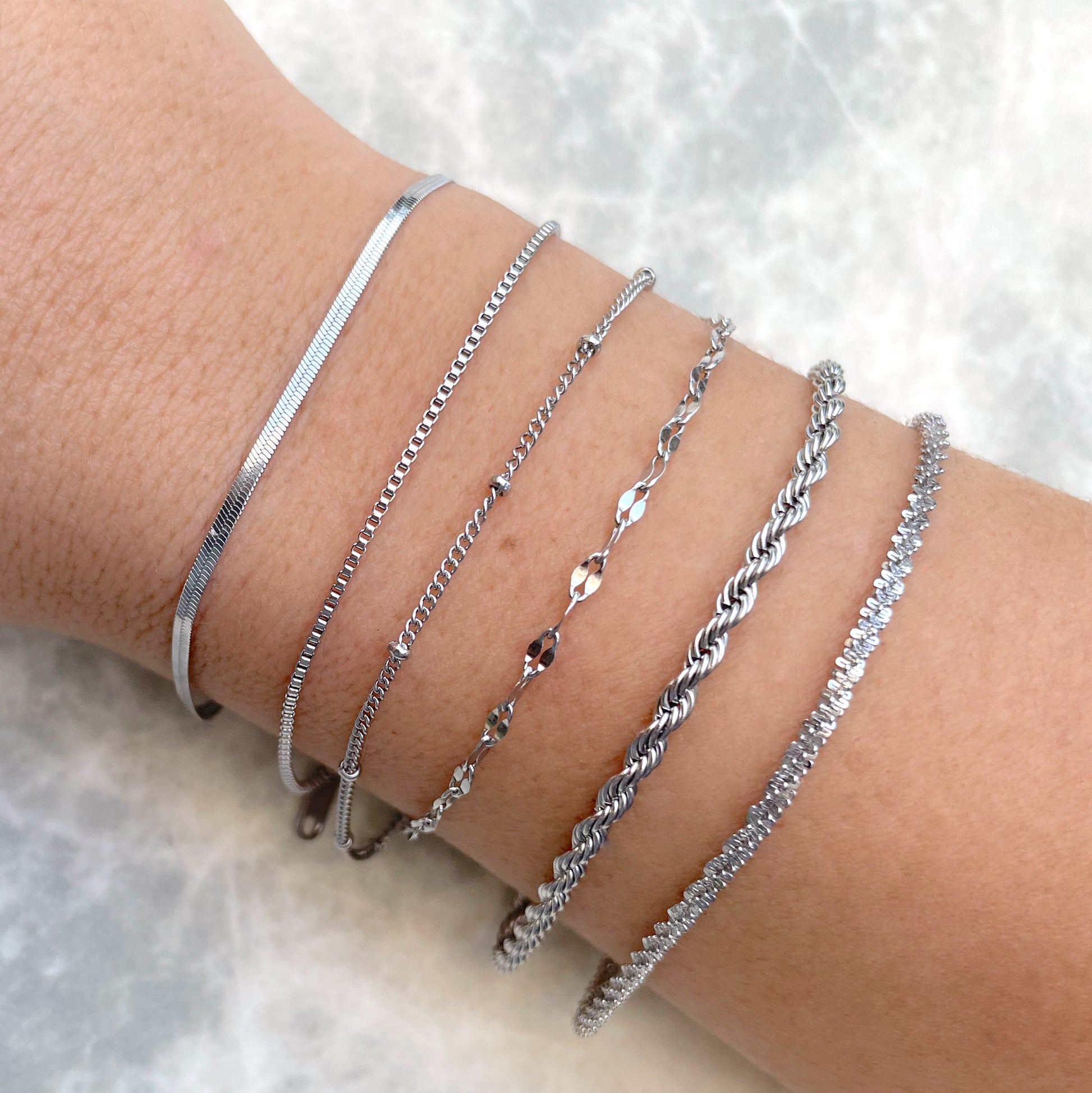 Close-up of a wrist wearing a dainty silver rope bracelet stacked with other stainless steel bracelets on a light background