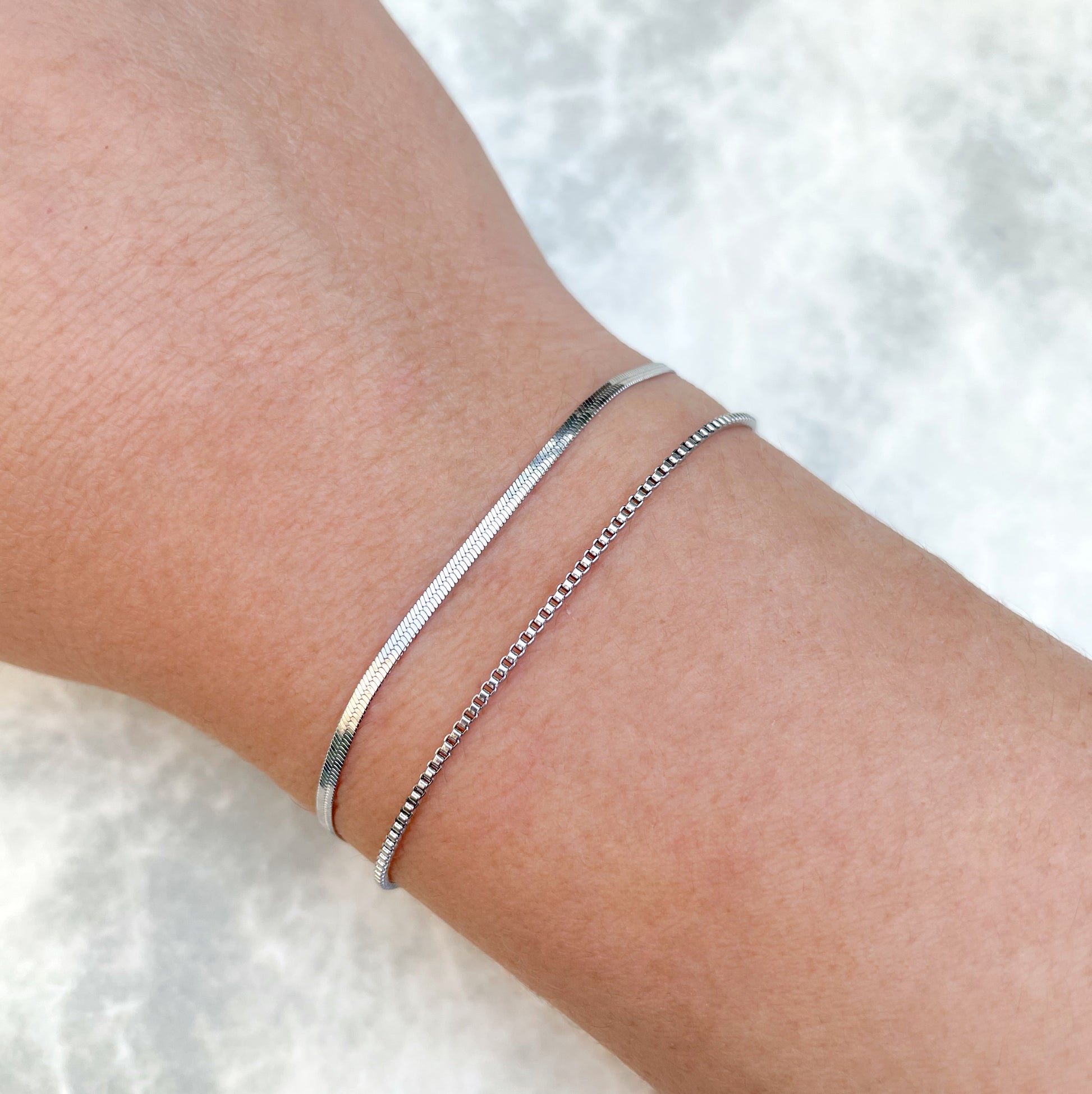 Silver box chain and herringbone bracelets stacked on a wrist against a white background