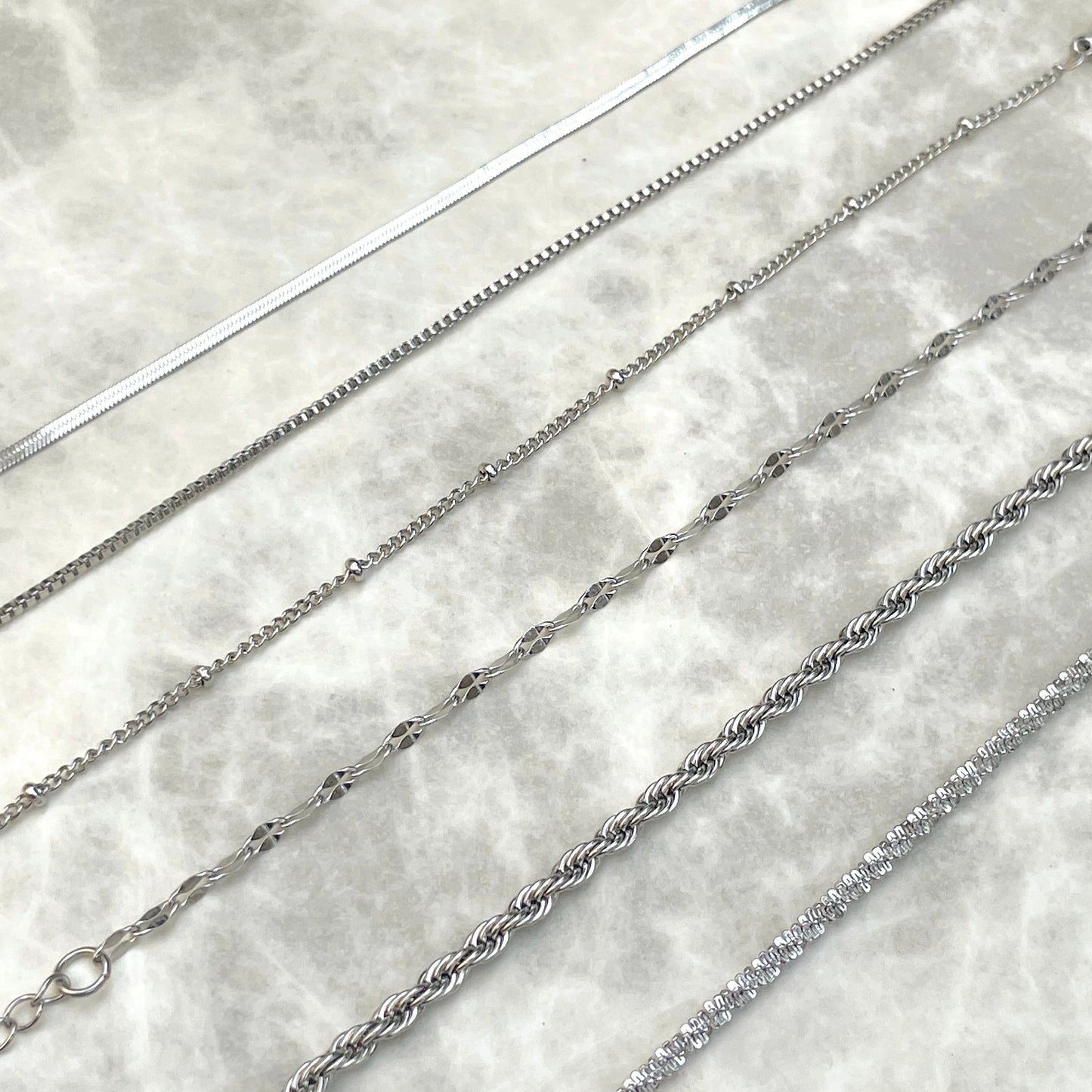 Six silver chain bracelets of different styles inclusing a 2mm herringbone, box, satellite, mariner, rope and sparkle rope, on a marble surface