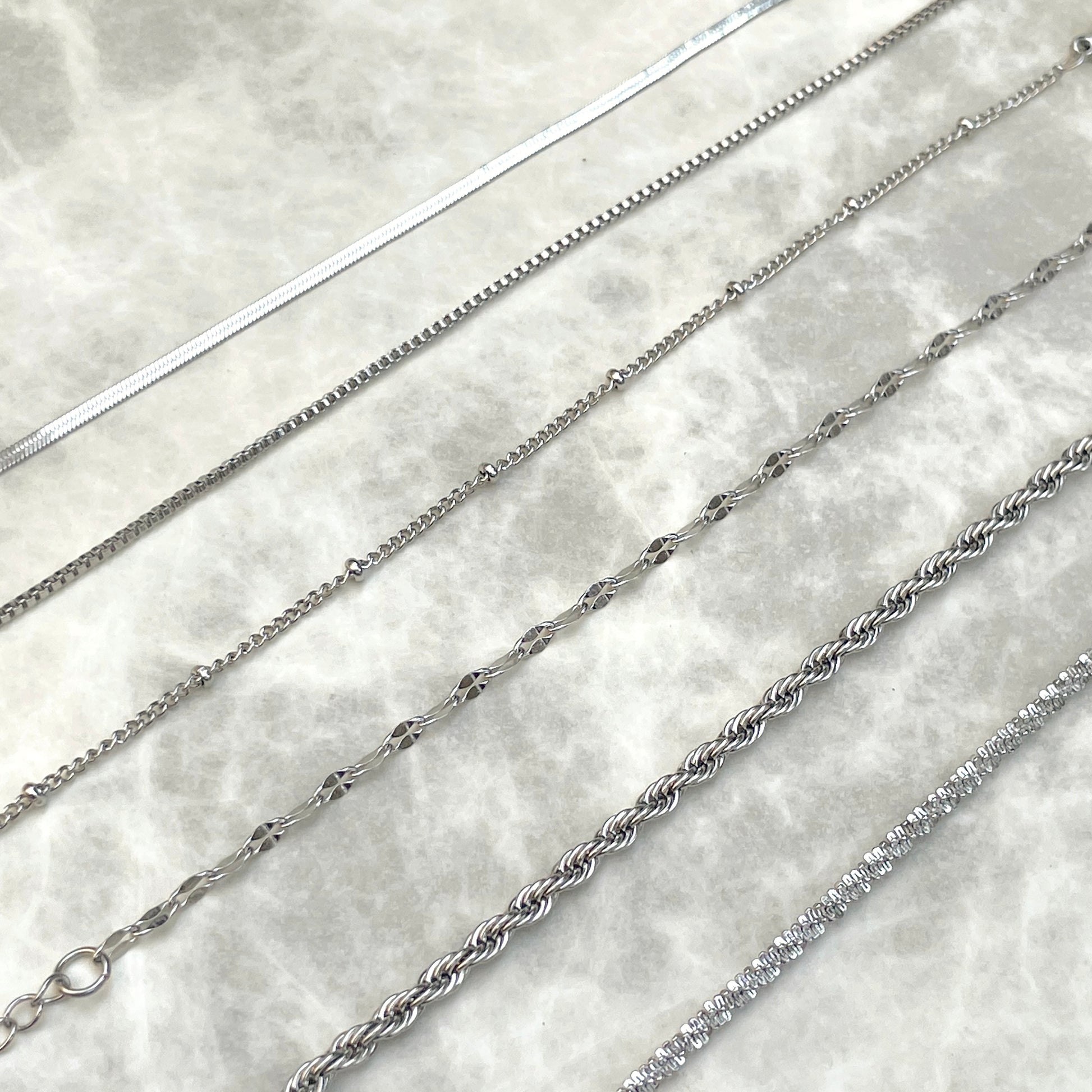 Six silver chain bracelets of different styles inclusing a 2mm herringbone, box, satellite, mariner, rope and sparkle rope, on a marble surface