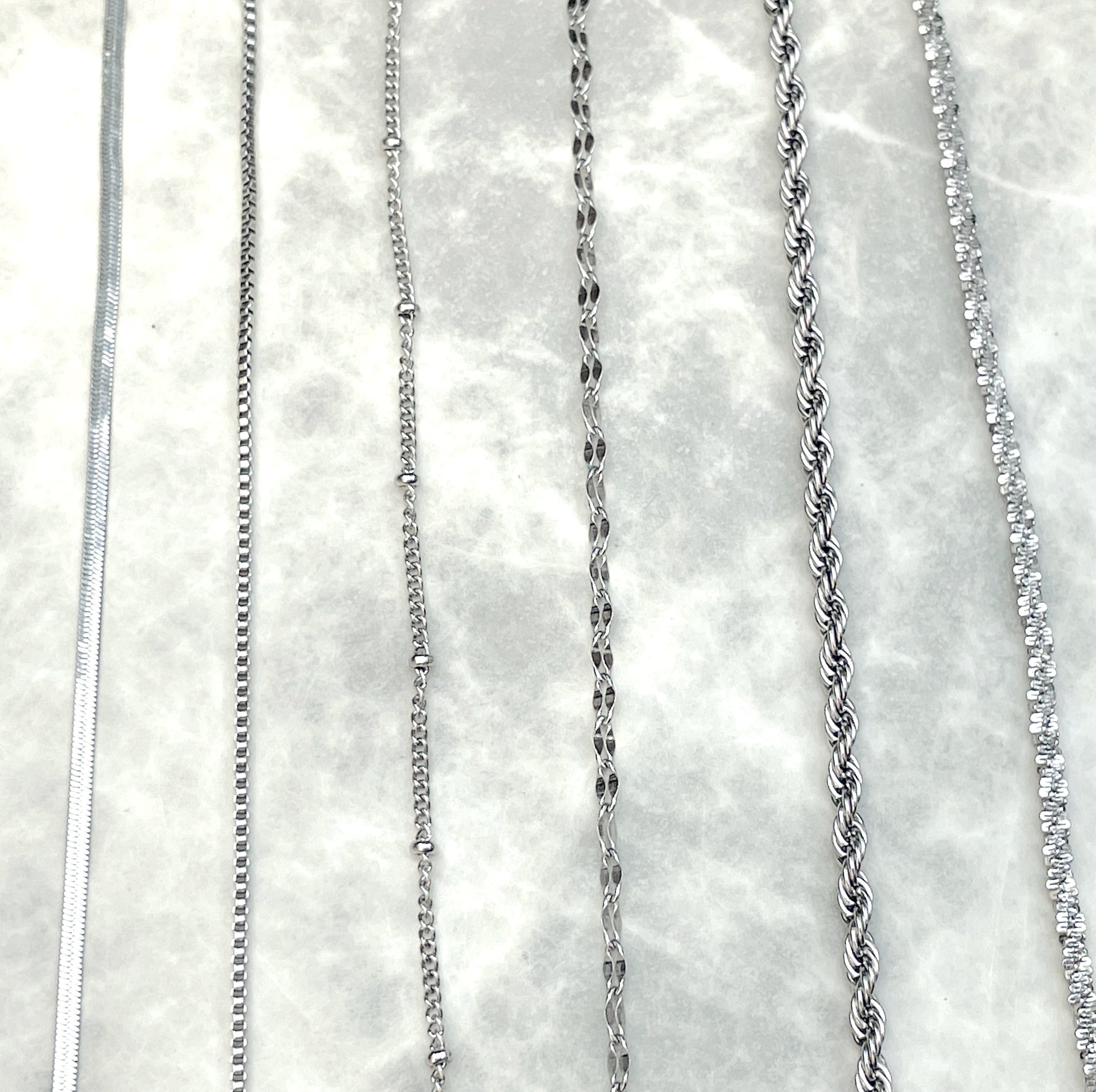 Top view of silver bracelet chain collection, vertically positioned on a textured white surface