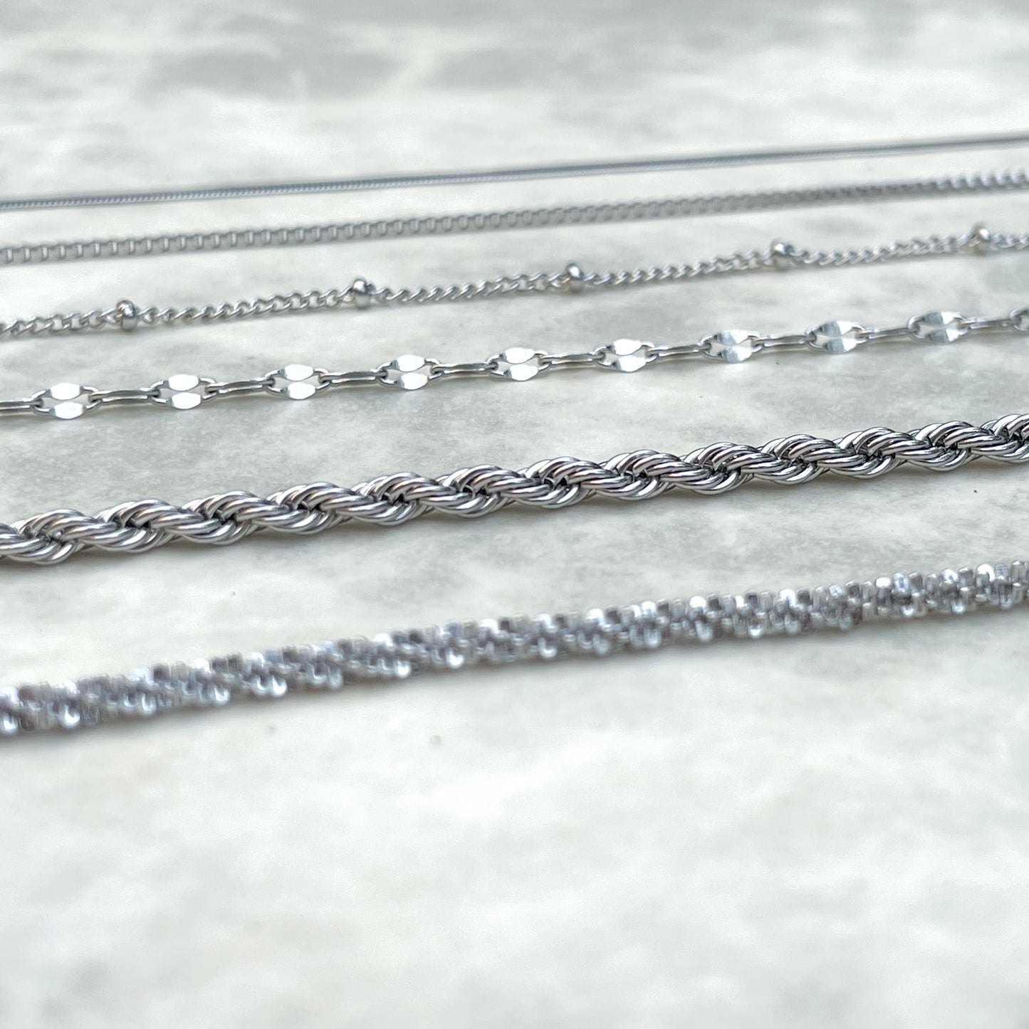 Close-up side view of silver chain bracelets on a marble surface, with focus being on rope and dainty mariner chains in the middle.