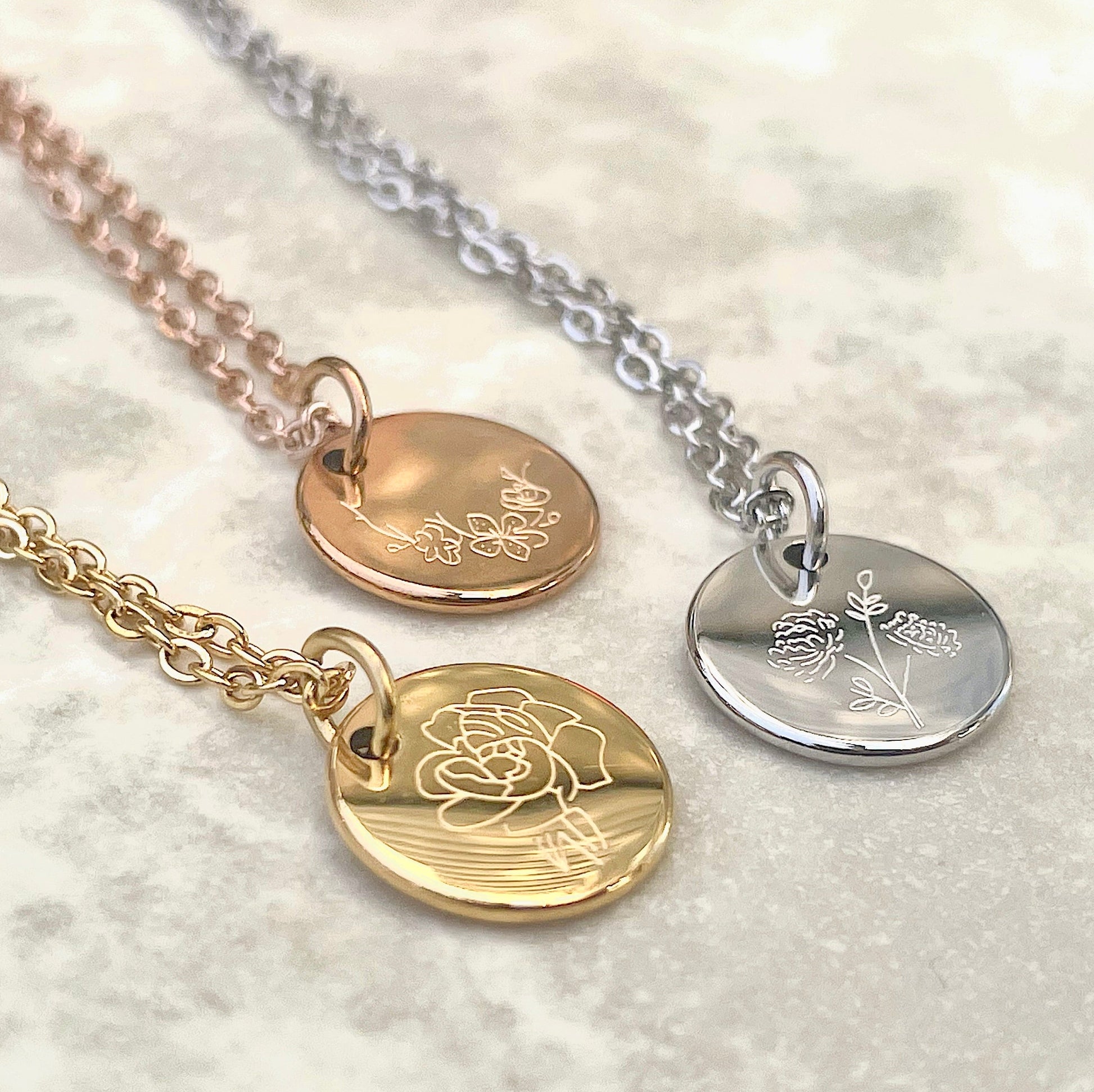 Three gold birth flower necklaces in rose gold, silver and gold with engraved pendants on a marble surface