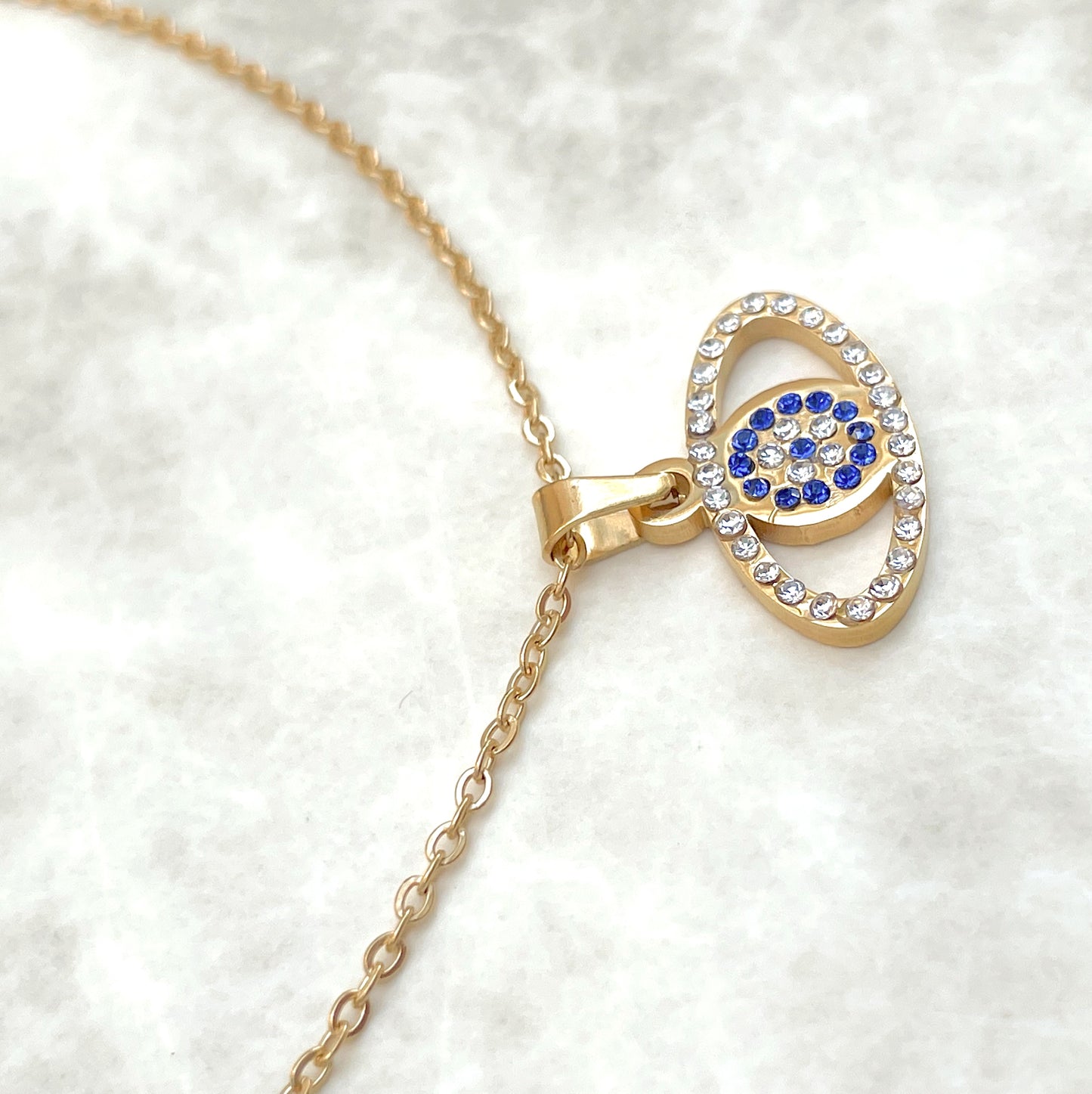 Close-up view of oval eye shaped pendant of the gold  evil eye necklace showcasing blue and clear CZ inlays on a white background