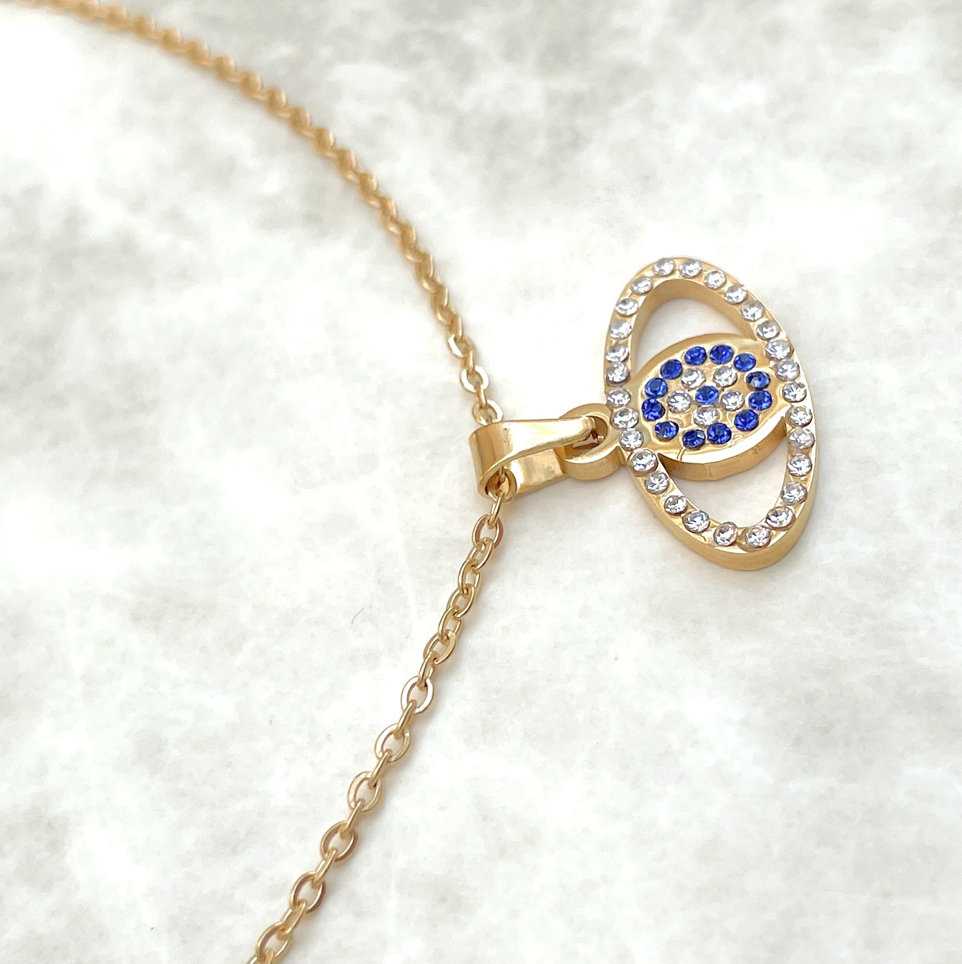 Close-up view of oval eye shaped pendant of the gold  evil eye necklace showcasing blue and clear CZ inlays on a white background