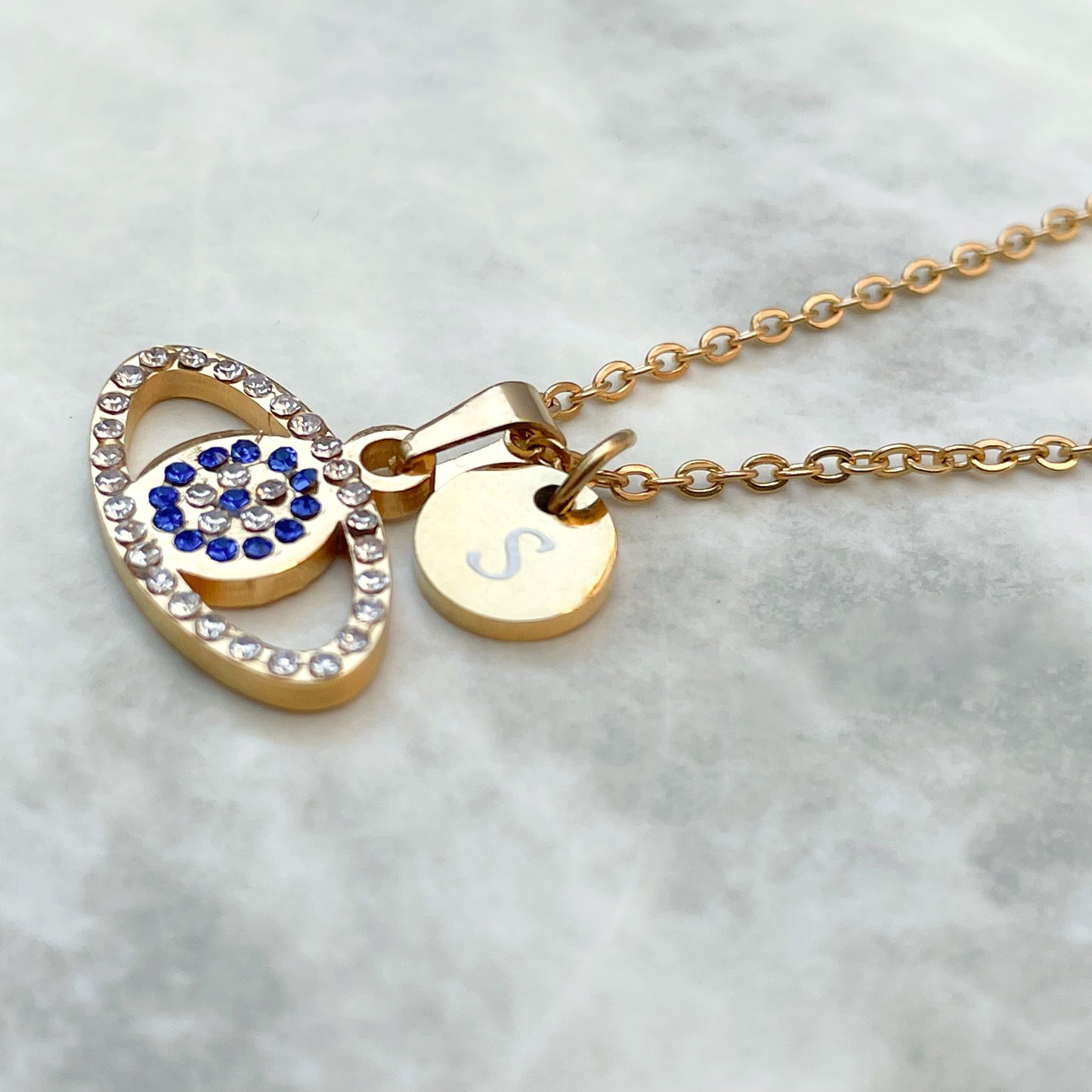 Gold eye necklace with an oval evil eye pendant and round initial disc with 'S' on a light gray background