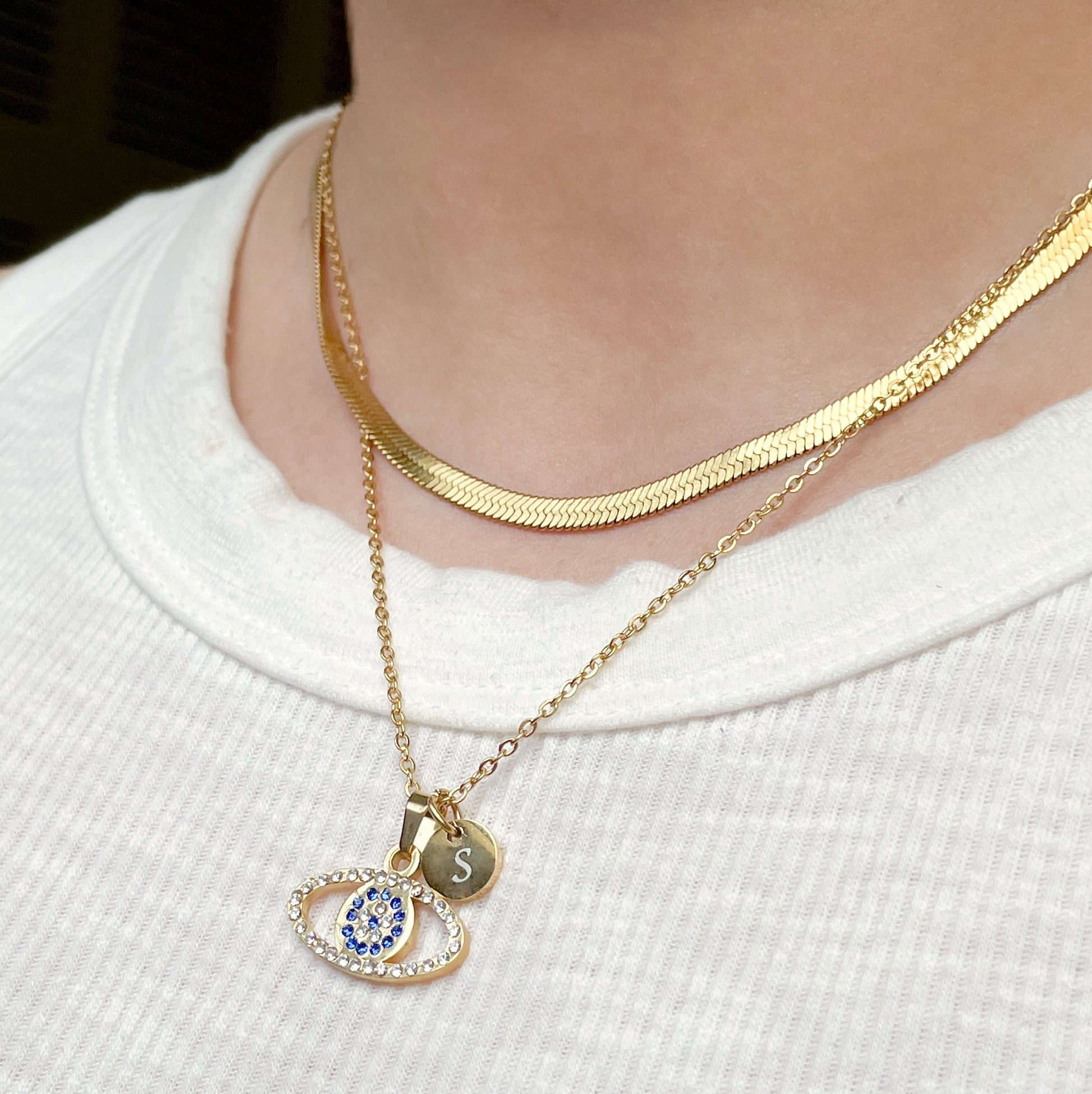 Gold necklace with an eye-shaped pendant and a small round initial charm worn by a person on a white background
