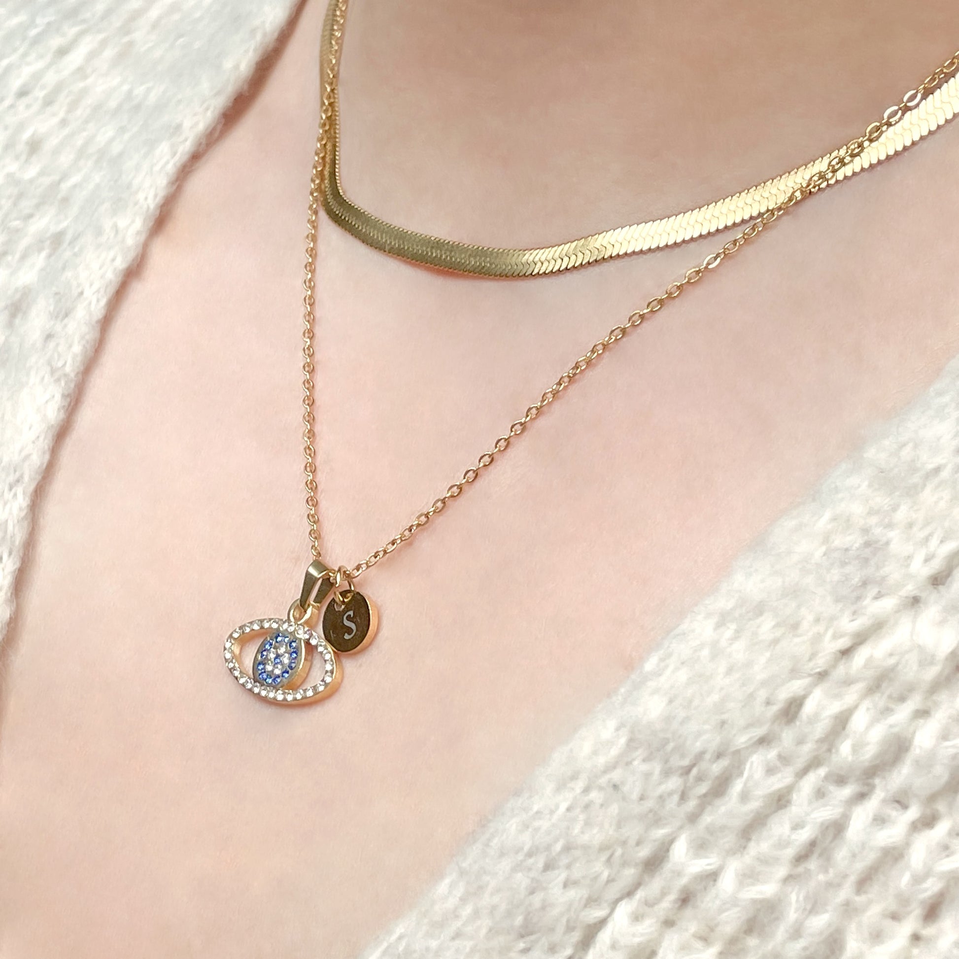 Gold protection necklace with an eye pendant paired with a 4mm herringbone choker on a person wearing a white textured garment.