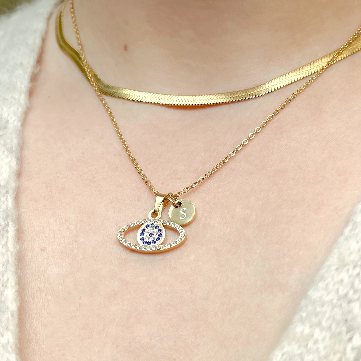Front view of gold necklace with an oval-shaped nazar pendant and small round disc with 'S' on a person's neck.