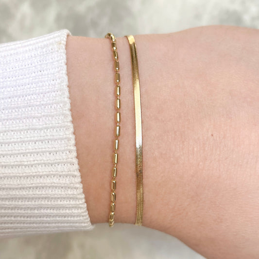 Layered gold bracelet with a dainty herringbone chain and oval link chain on a wrist with a white sleeve