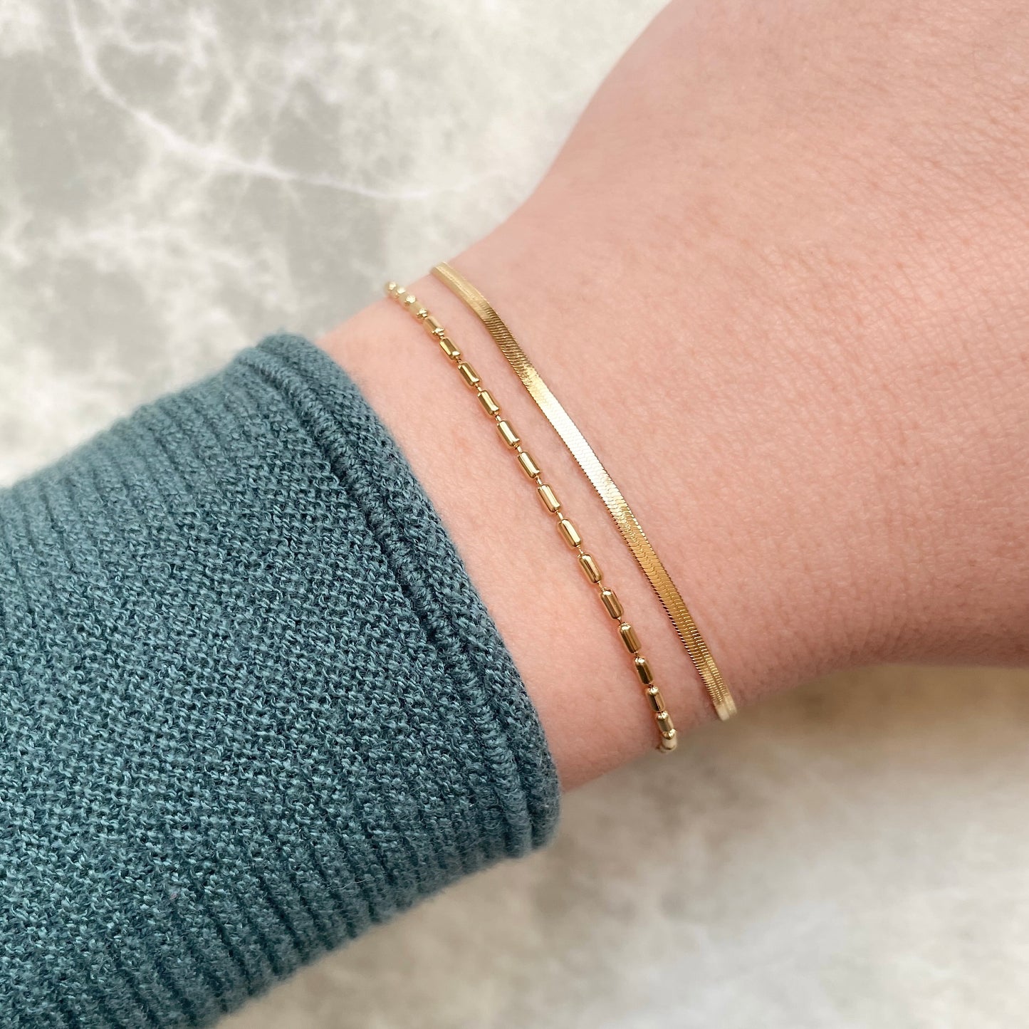 Angled top view of  dainty gold bracelet set on a wrist with a textured sleeve against a neutral background