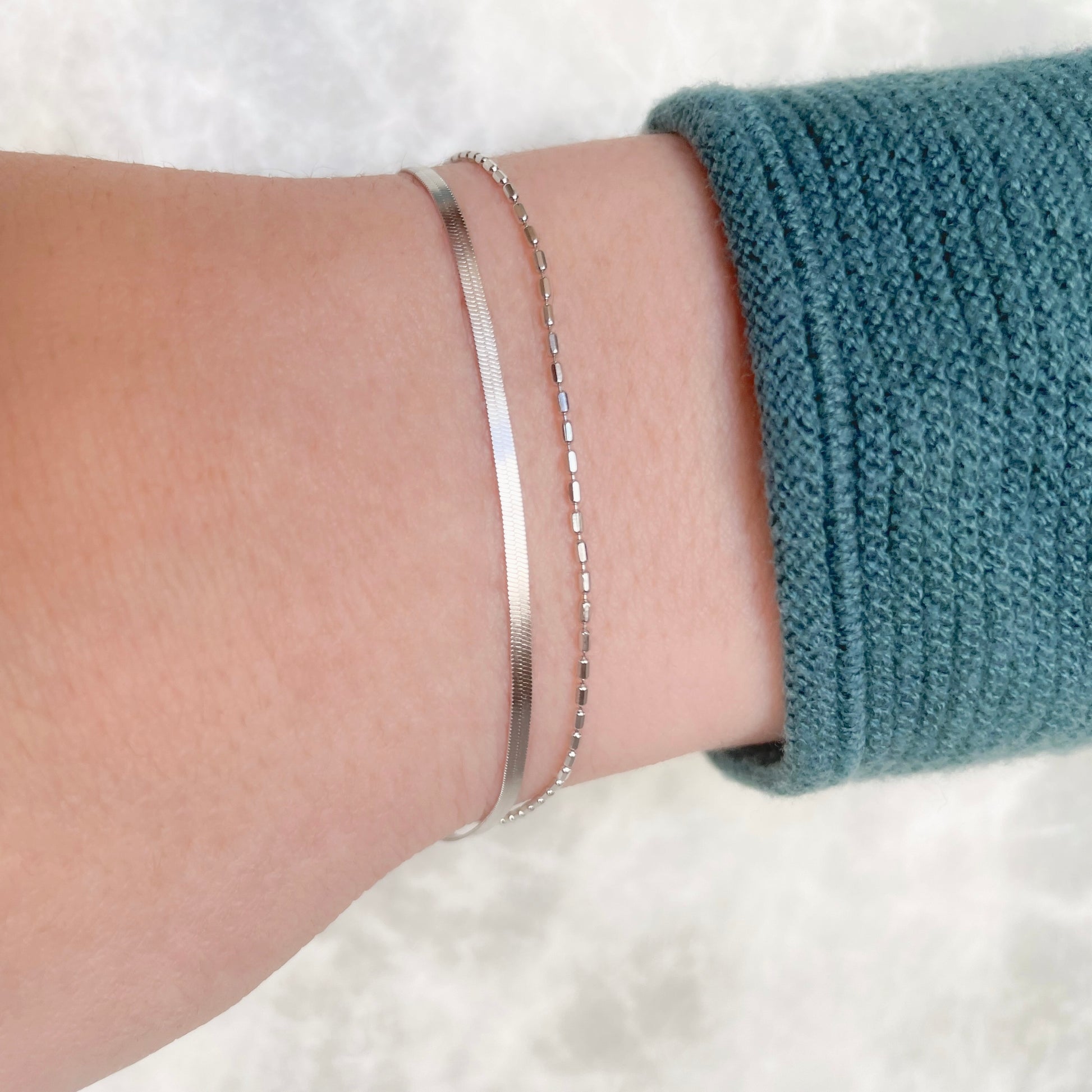 Close-up of a wrist with a silver herringbone and ball chain bracelet on a white background