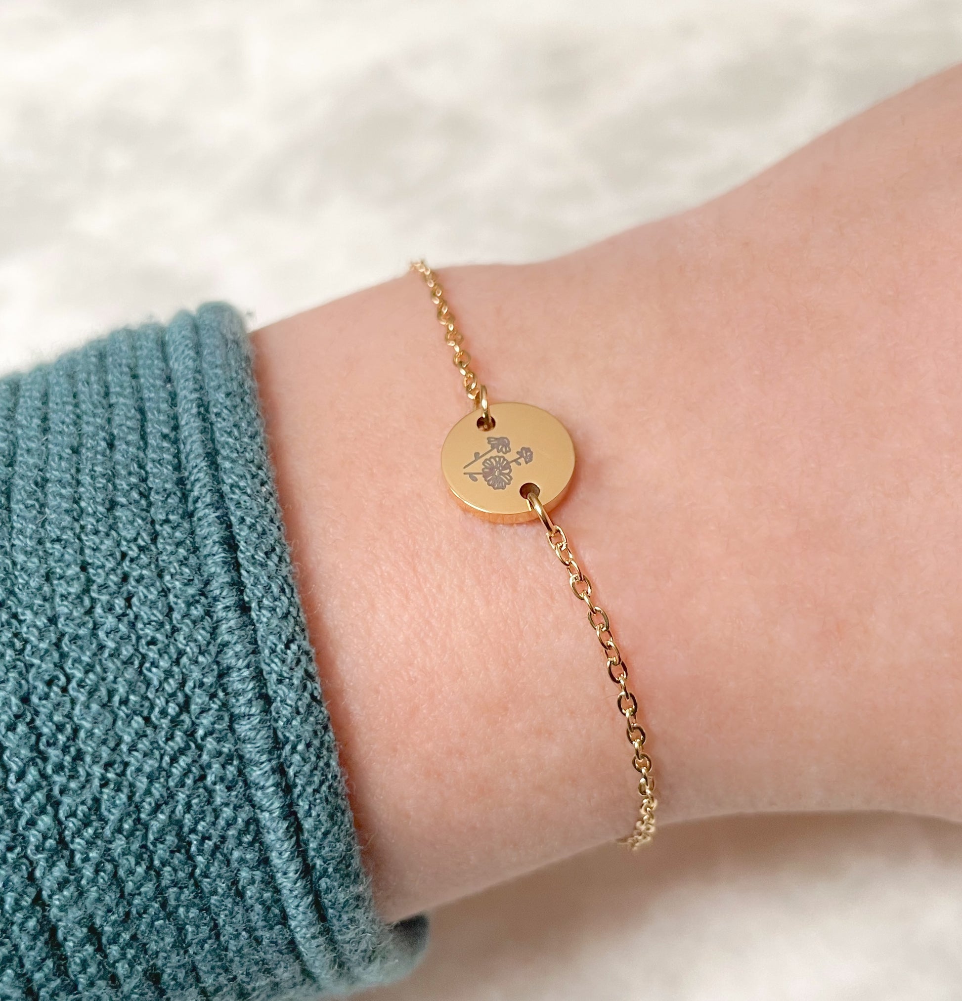 Side view of gold bracelet with a daisy engraved round charm  on a wrist, wearing a teal sleeve