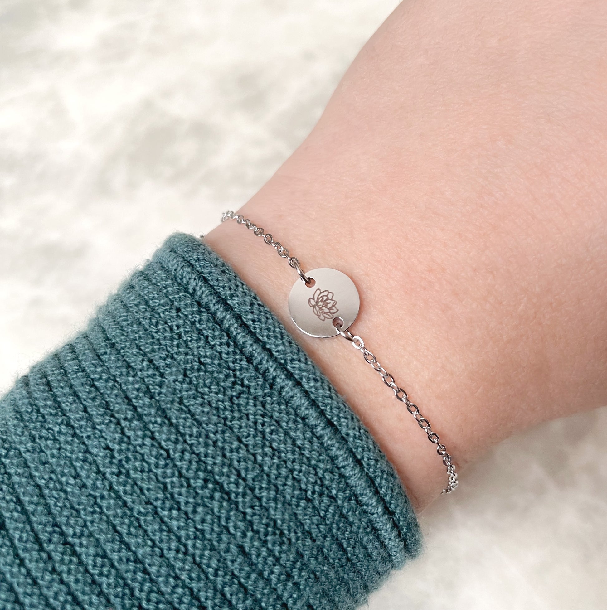 Silver bracelet with a lotus flower engraved round charm on a wrist wearing a teal sleeve against a white background