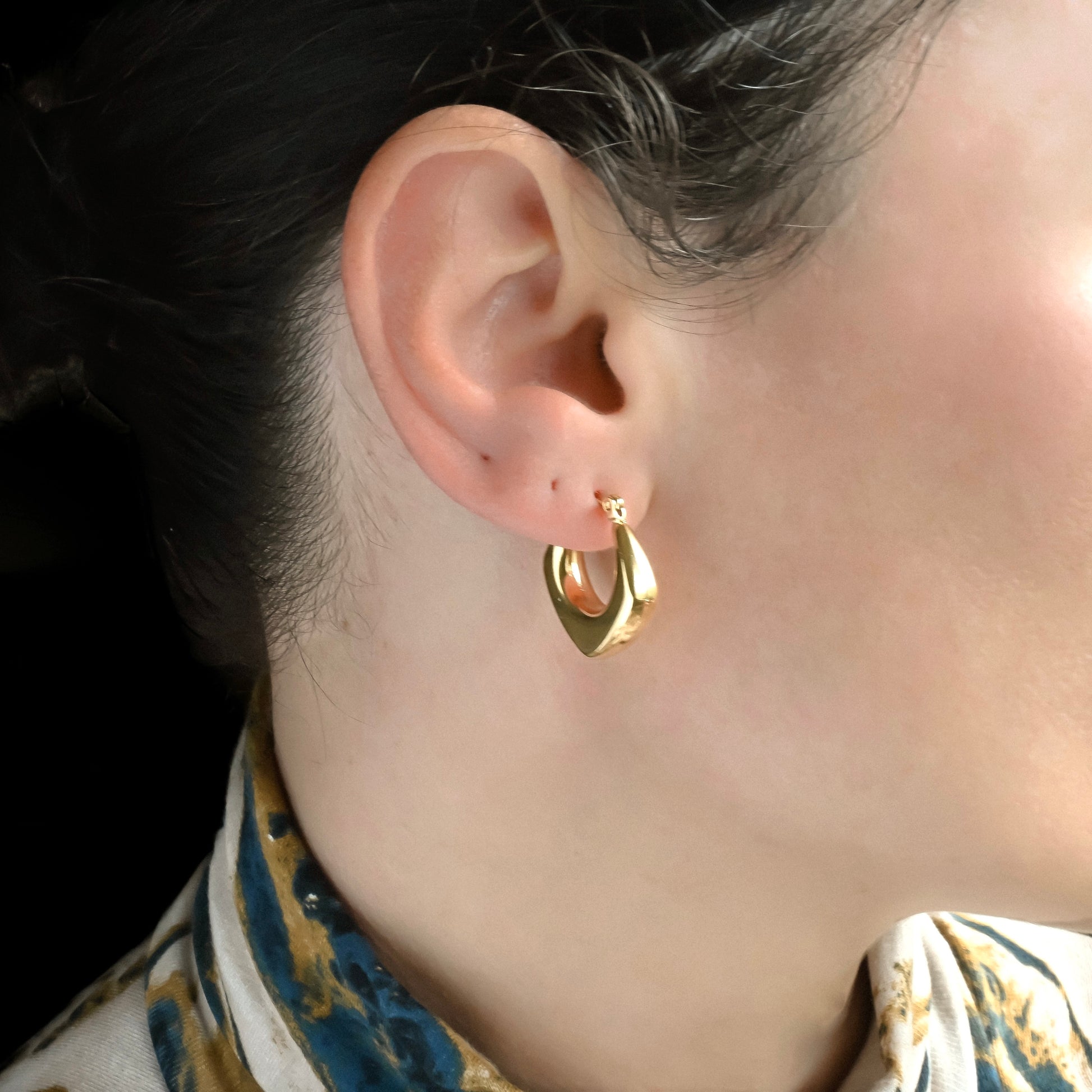 Gold rhombus hoop earrings worn by a person with a blurred background