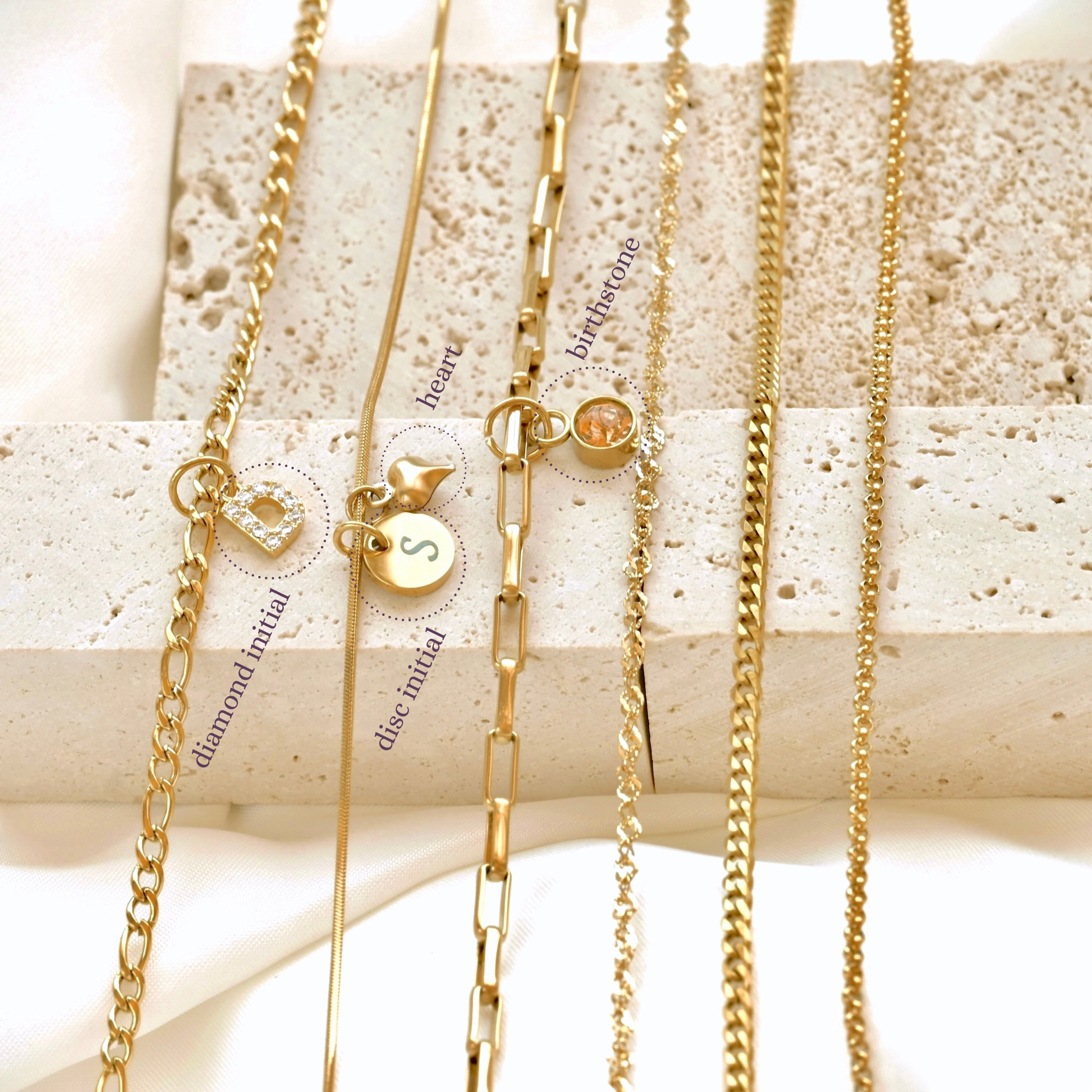 Dainty Gold necklaces with various charms( diamond initial, heart, round initial and birthstone) on a textured surface