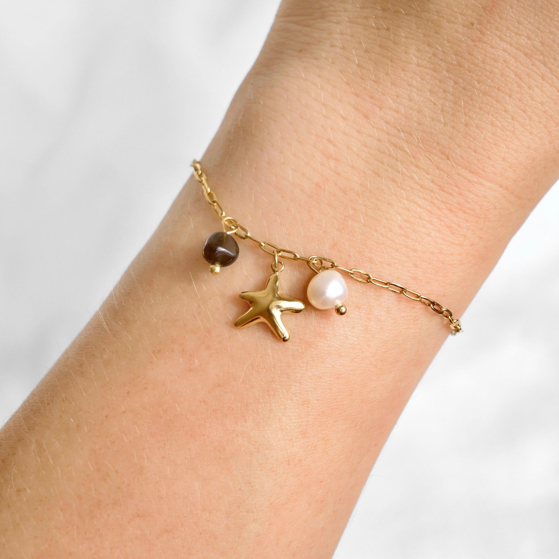 A delicate gold paperclip chain bracelet with a starfish charm, a white pearl charm, and a black stone charm, shown worn on a person's wrist.