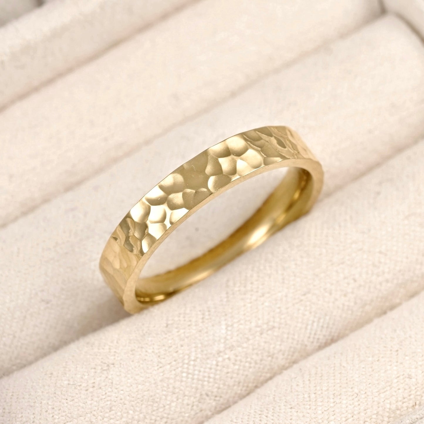 A gold band ring with a hammered texture, resting on a cream-colored fabric ring box.