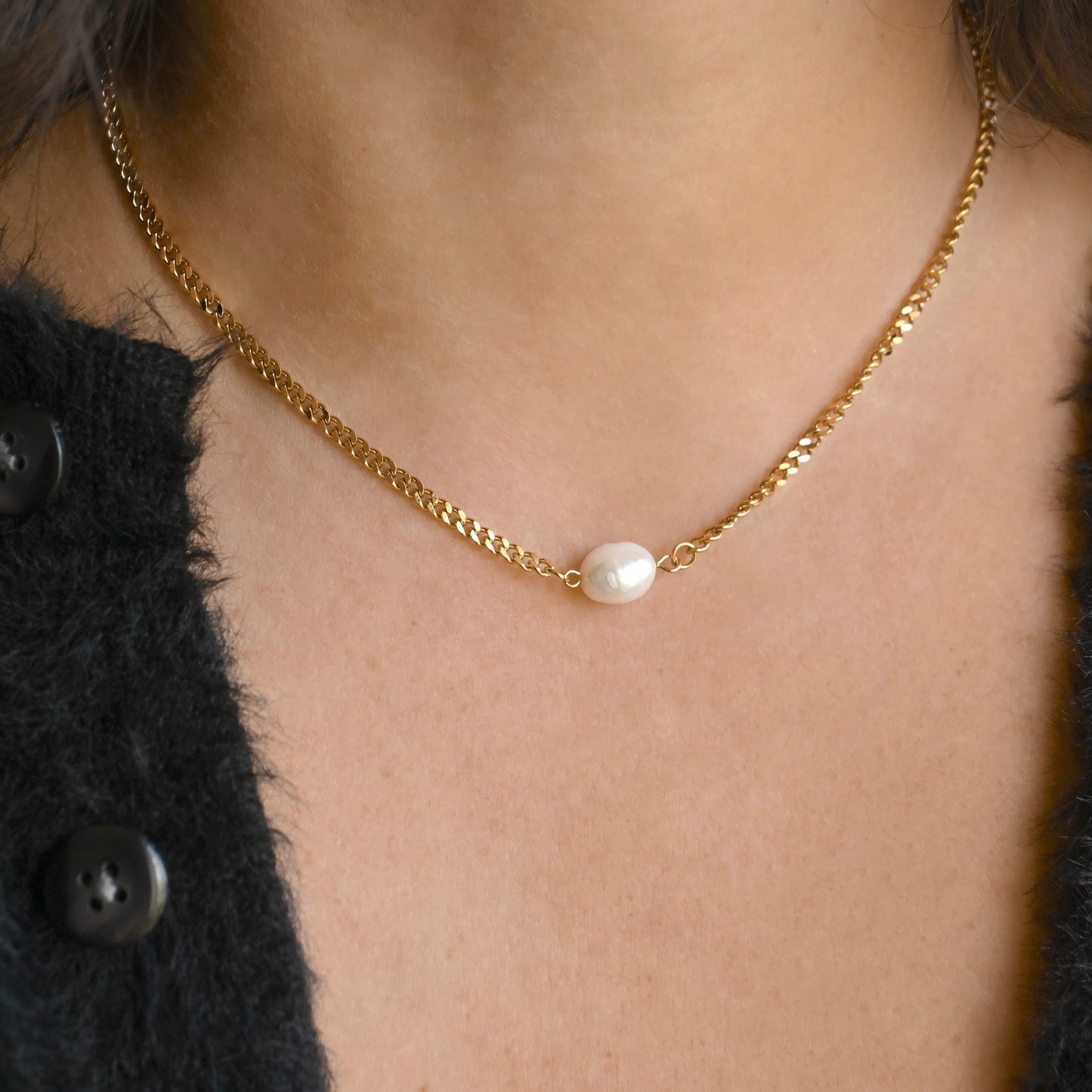 A close-up of a person's neck and chest wearing a delicate gold chain necklace with a single white baroque pearl pendant on a 3mm curb chain, styled with a black textured cardigan.