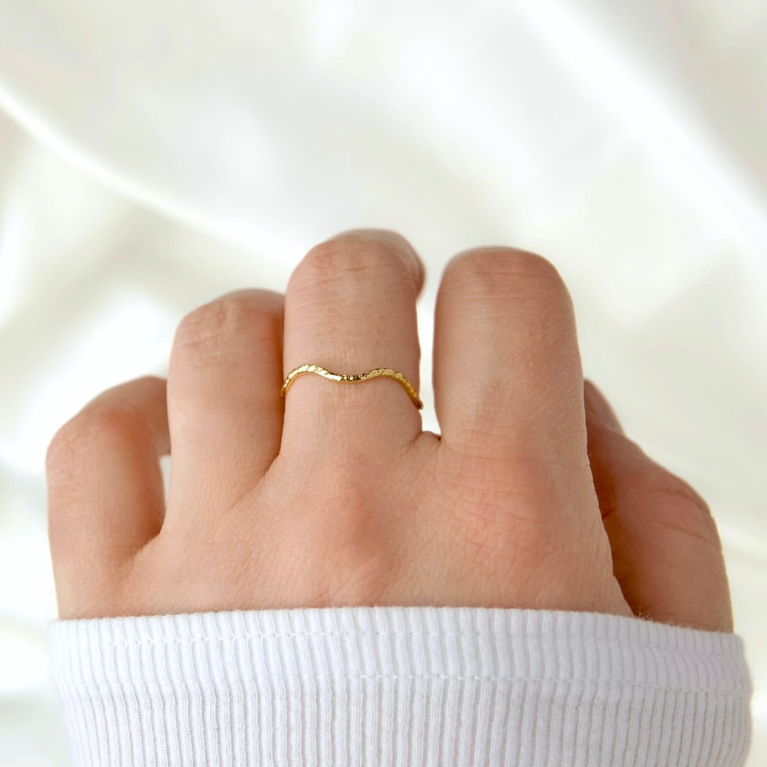 A delicate, thin gold ring with a wavy, organic texture worn on a person's ring finger, shown against a soft, draped white fabric background.